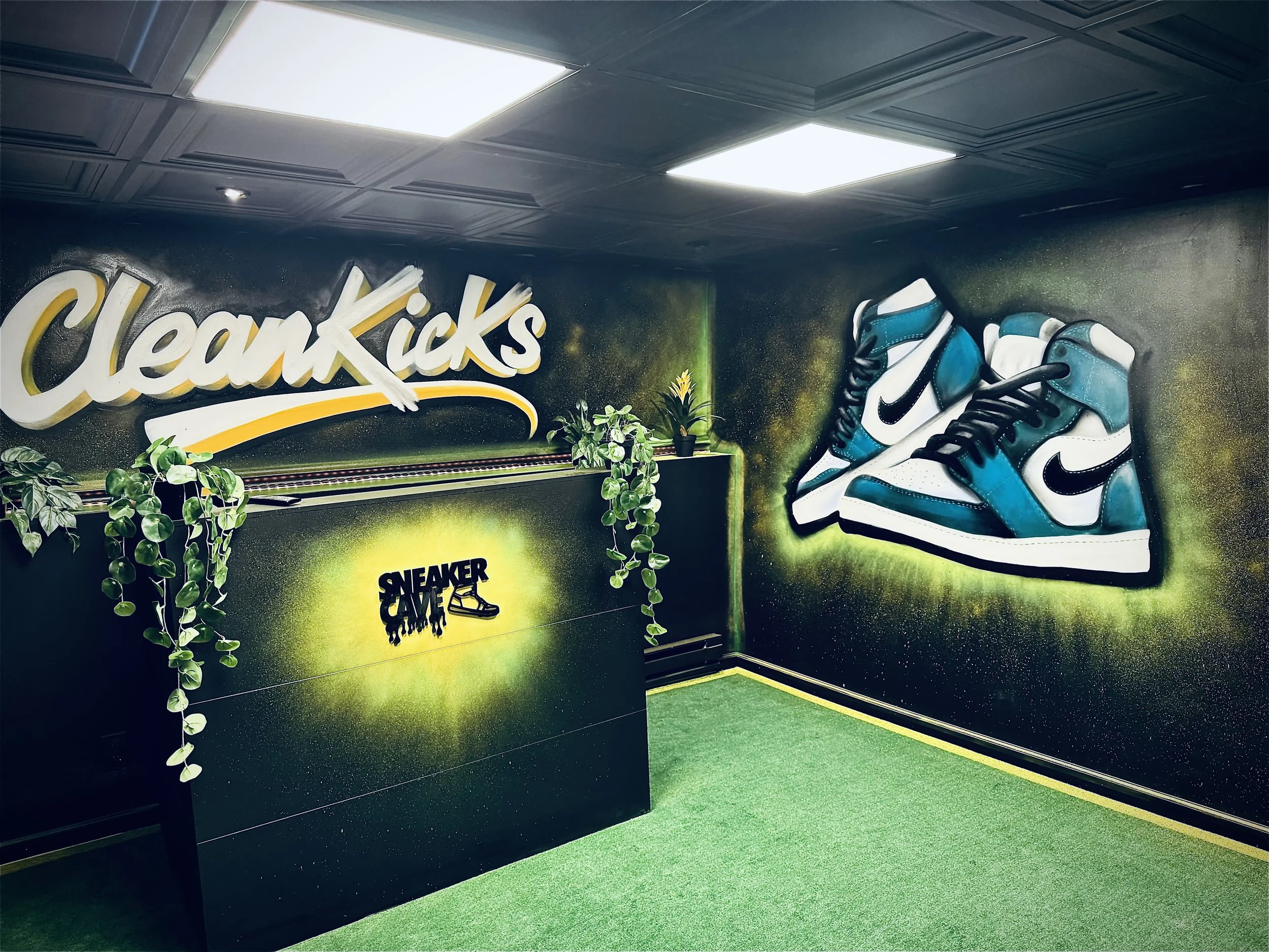 Interior of a sneaker store with a wall decorated with a neon "Sneaker Cave" sign, sneakers on display, artificial plants, and green turf flooring.