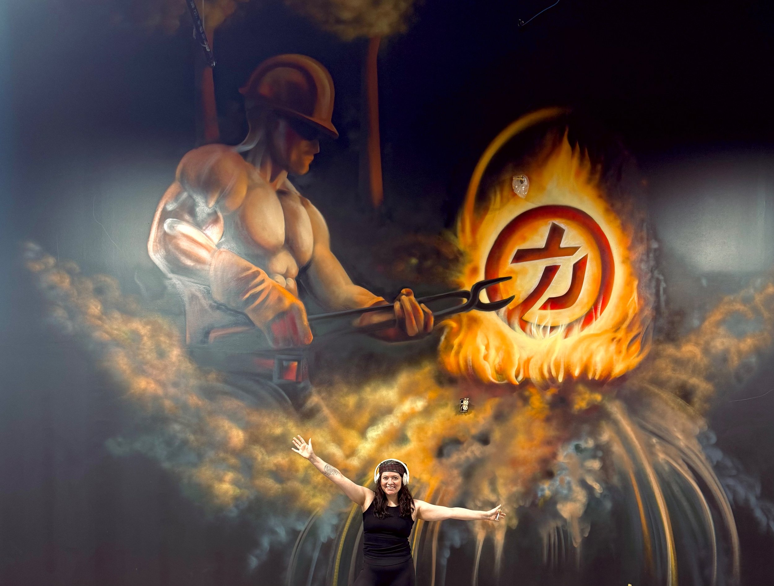 A woman smiling and raising her arms, wearing headphones, with a background resembling a fiery explosion illuminating a symbol on the wall.