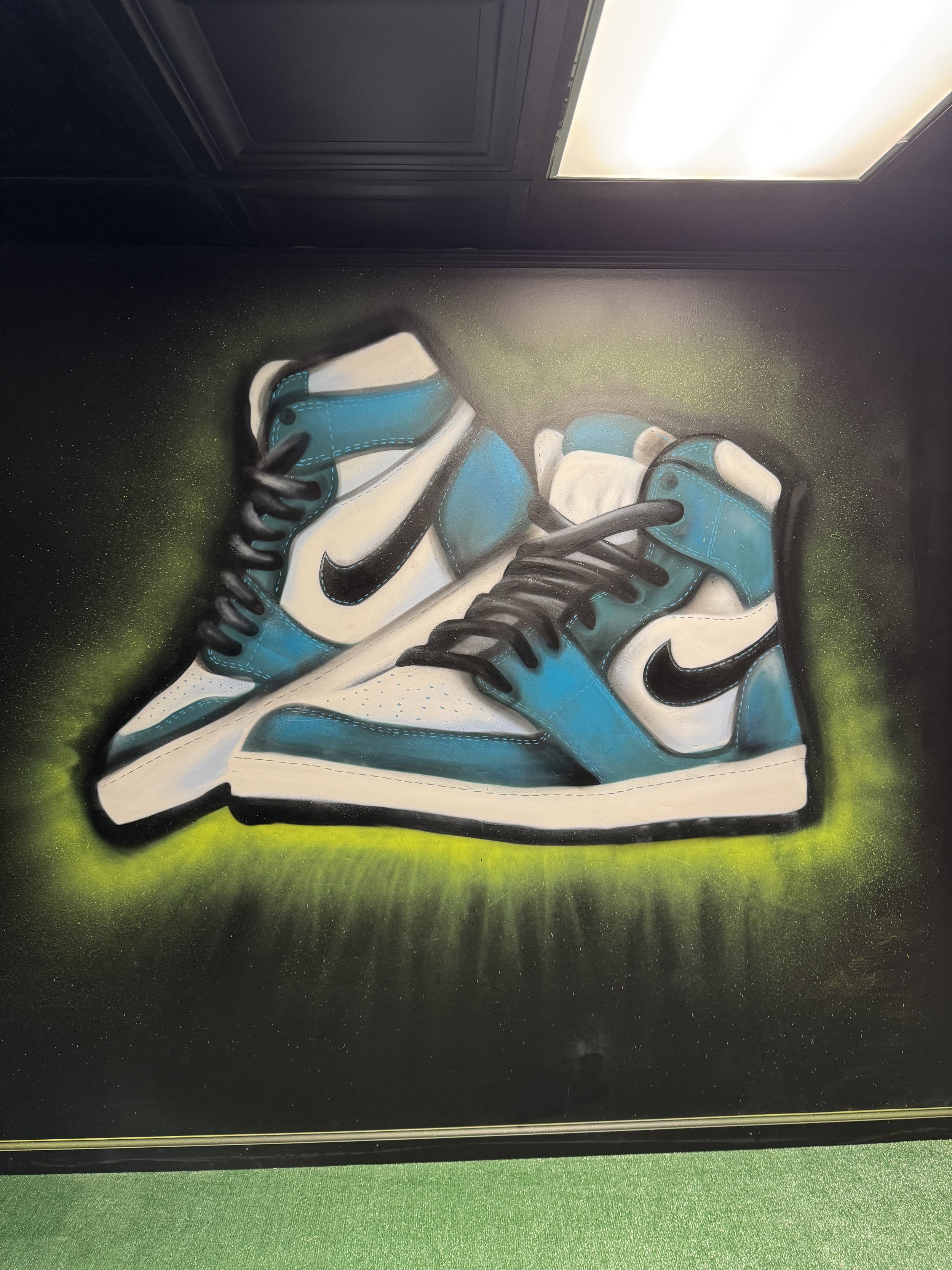 A painting of a pair of Nike sneakers with blue, white, and black colors, set against a black background with green spray paint accents.