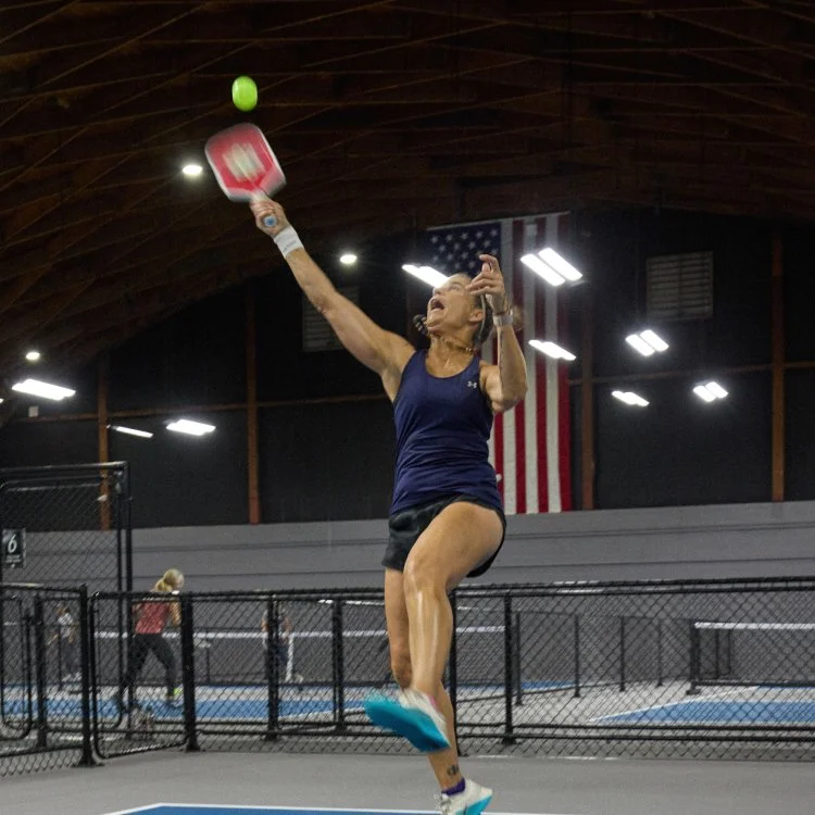Woman playing pickleball on an indoor court, jumping to hit a green ball with a pink paddle, with an American flag in the background.
