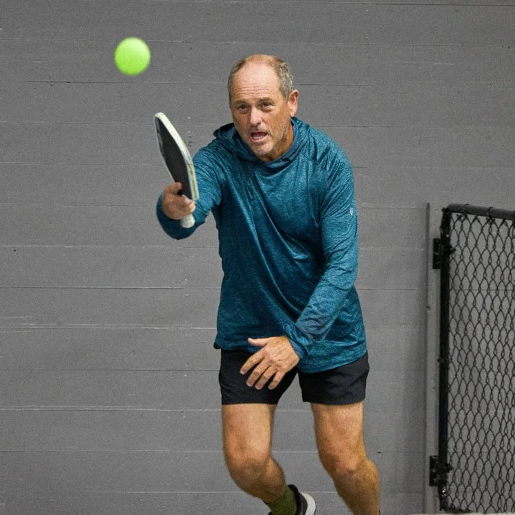 Man wearing blue long-sleeve shirt and black shorts playing tennis indoors against a gray wall.