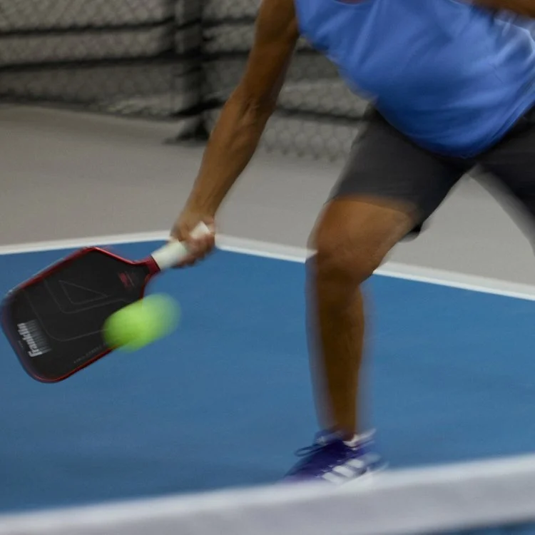 Person in athletic clothing hitting a tennis ball with a paddle on a blue court.