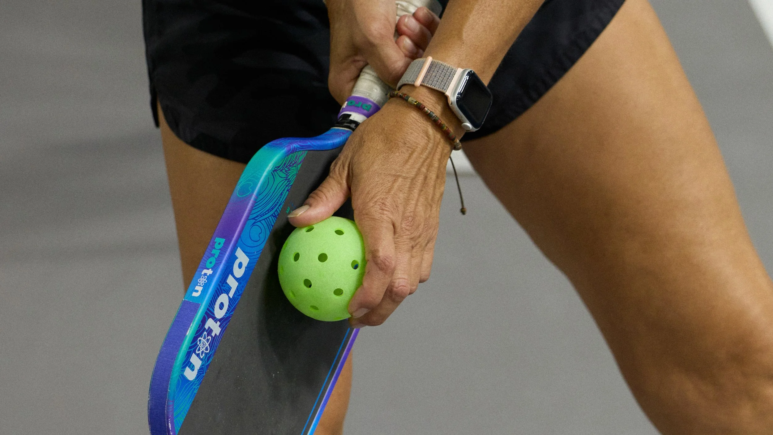 A person holding a green perforated ball with their left hand, a colorful paddle in their right hand, wearing a watch and a beaded bracelet, with gray background.