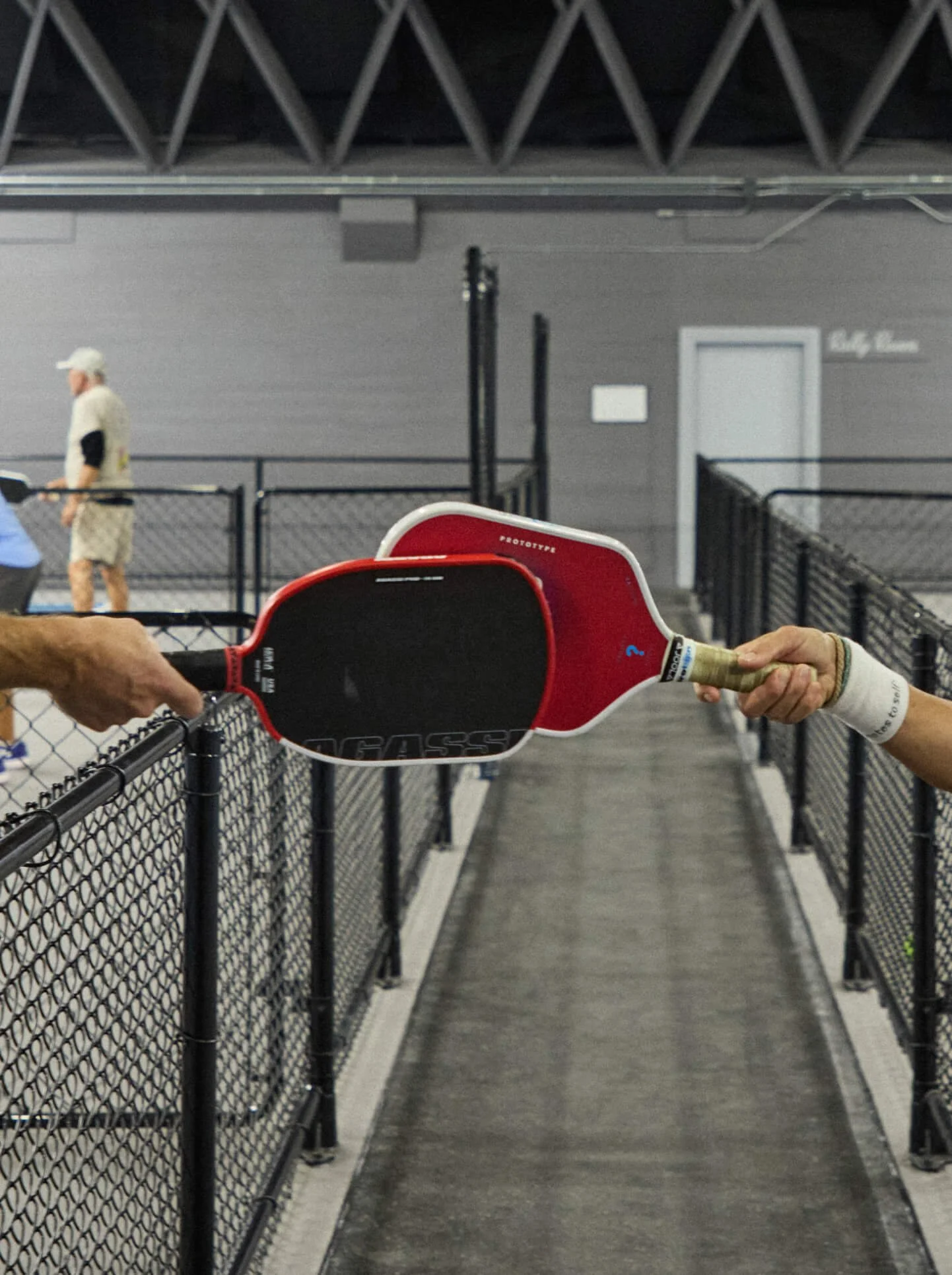 Close-up of two padel rackets, one red and one black, being held by players at the net, with a netted walkway and a man in beige shorts and a white hat in the background at an indoor sports facility.