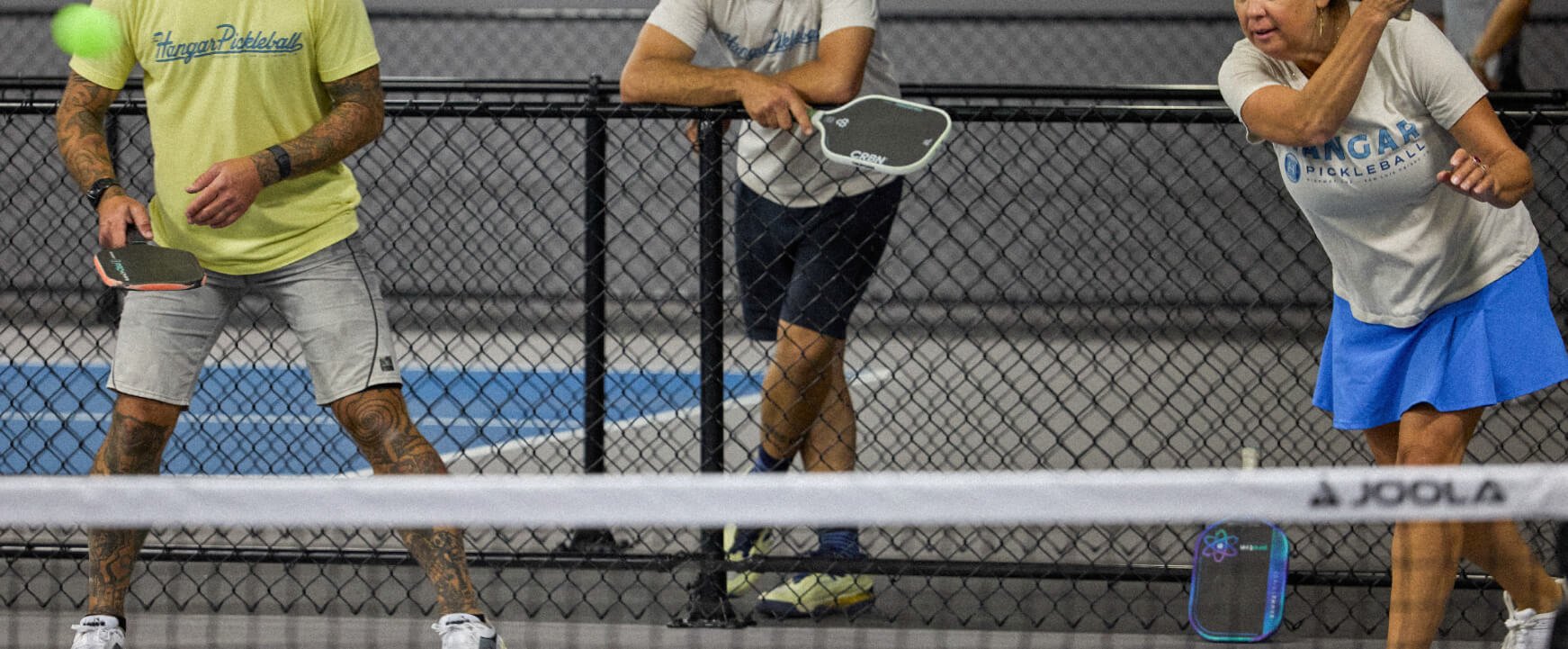 Three people playing pickleball inside an indoor court, two women and one man, with paddles and a green pickleball visible.