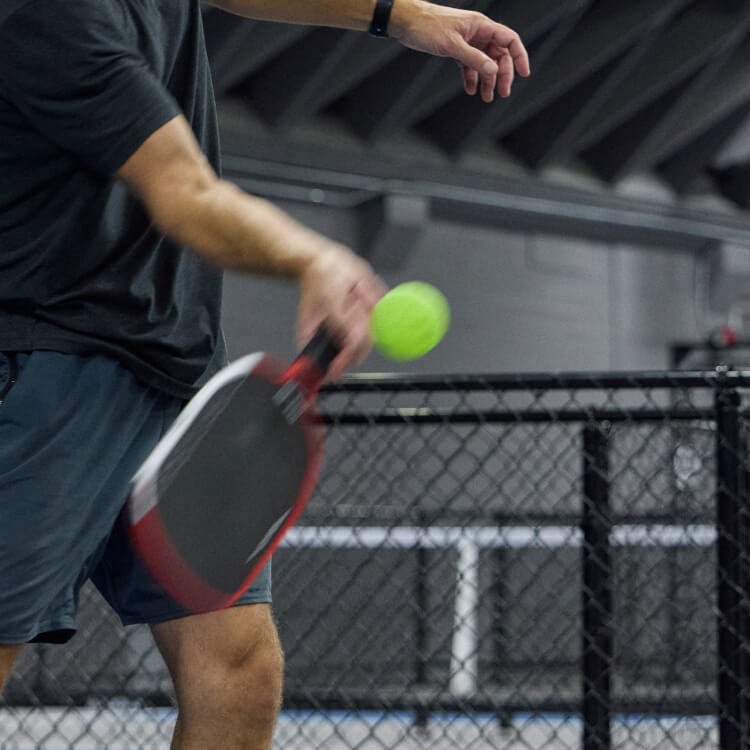 A person is playing pickleball indoors, holding a paddle and hitting a green pickleball.