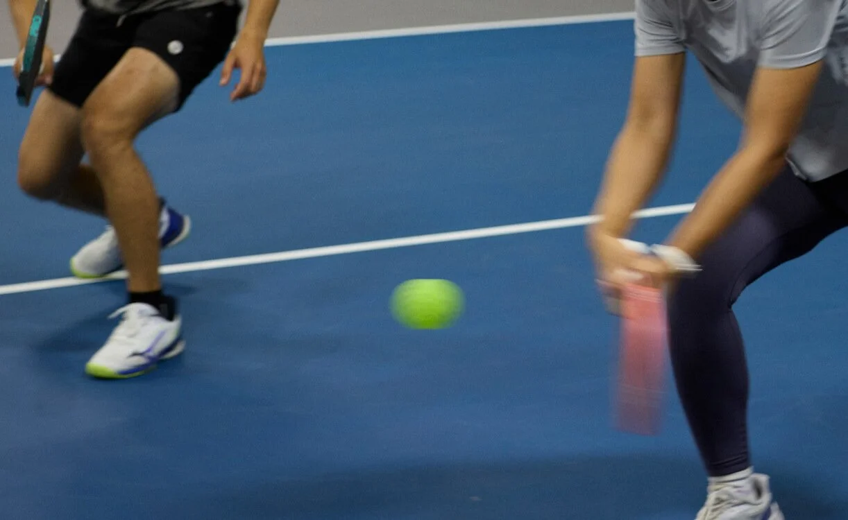 Two people playing tennis indoors with a bright green tennis ball between them on a blue court.