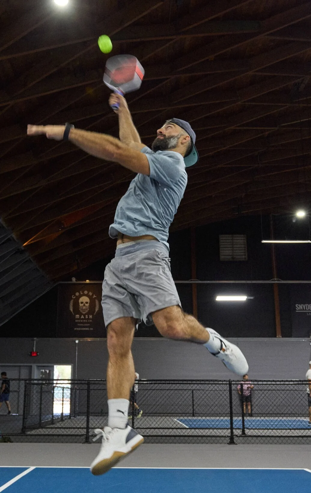 A man playing pickleball in an indoor gym, jumping to hit a green pickleball with a paddle.