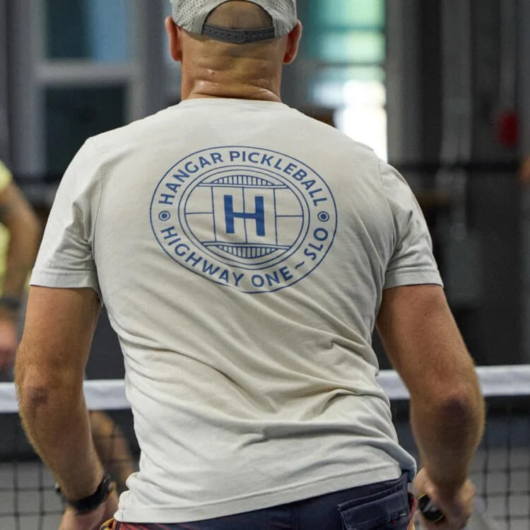 Person wearing a gray shirt with a logo on the back that reads 'HANGAR PICKLEBALL HIGHWAY ONE-0TS' at an indoor pickleball court.