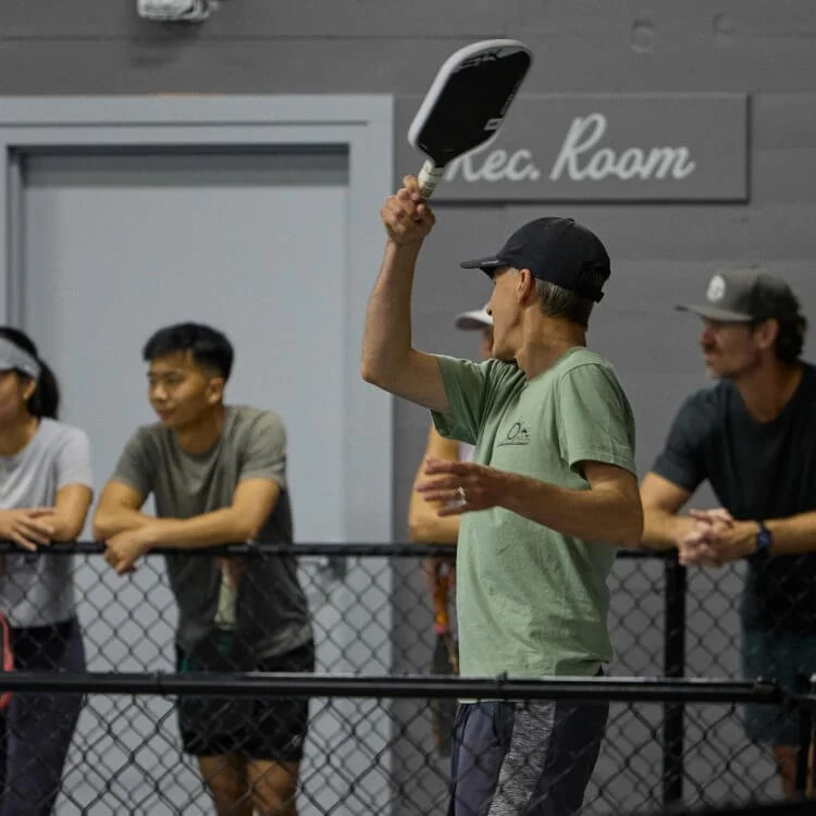 Group of people playing pickleball indoors, with one woman holding a paddle and preparing to serve.