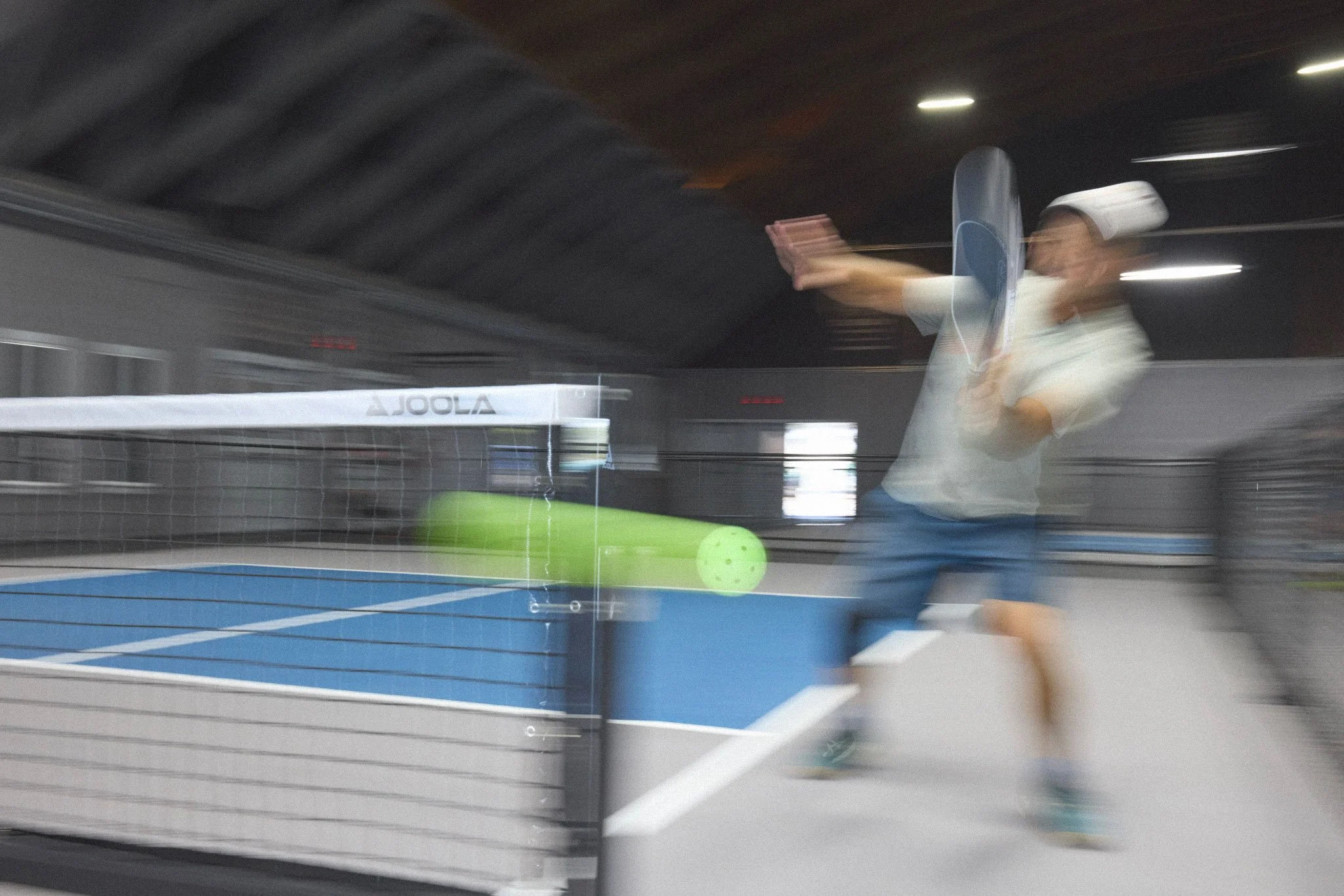 A woman playing table tennis indoors. The image is blurry due to motion, showing her in action with a paddle, with a green ball visible on the table.