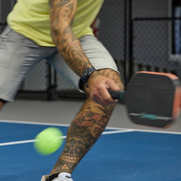 Person with tattoos hitting a tennis ball with a paddle on a blue court.
