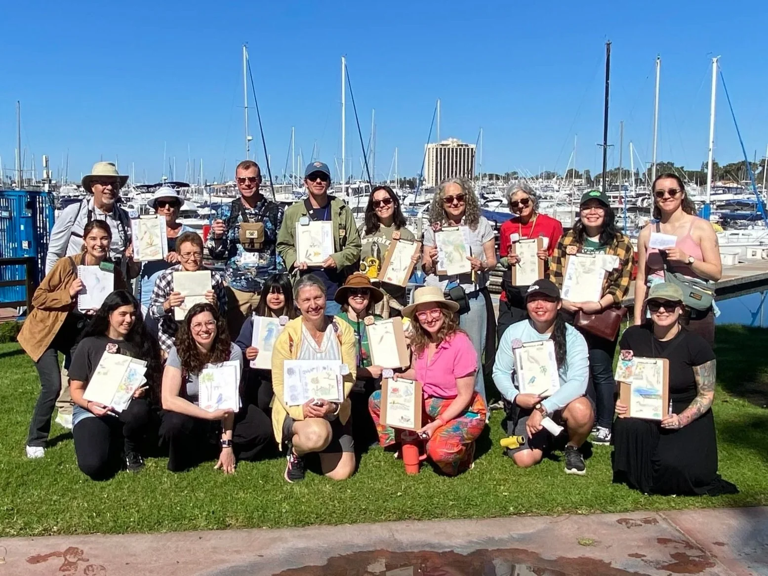 A large group of people smiling and holding up drawings or paintings, standing on a grassy area near a marina filled with boats and yachts under a clear blue sky.