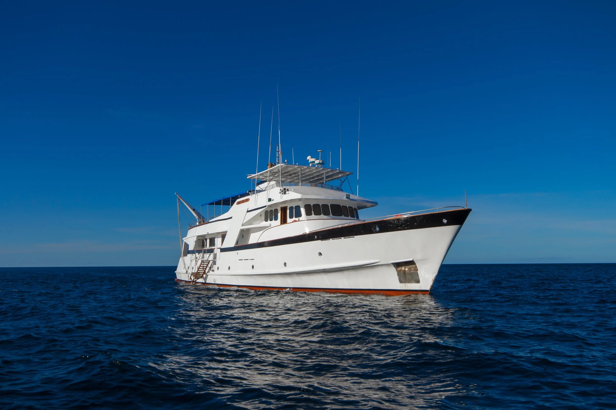 A large white yacht with antennas and radar on top floats on the blue ocean under a clear blue sky.