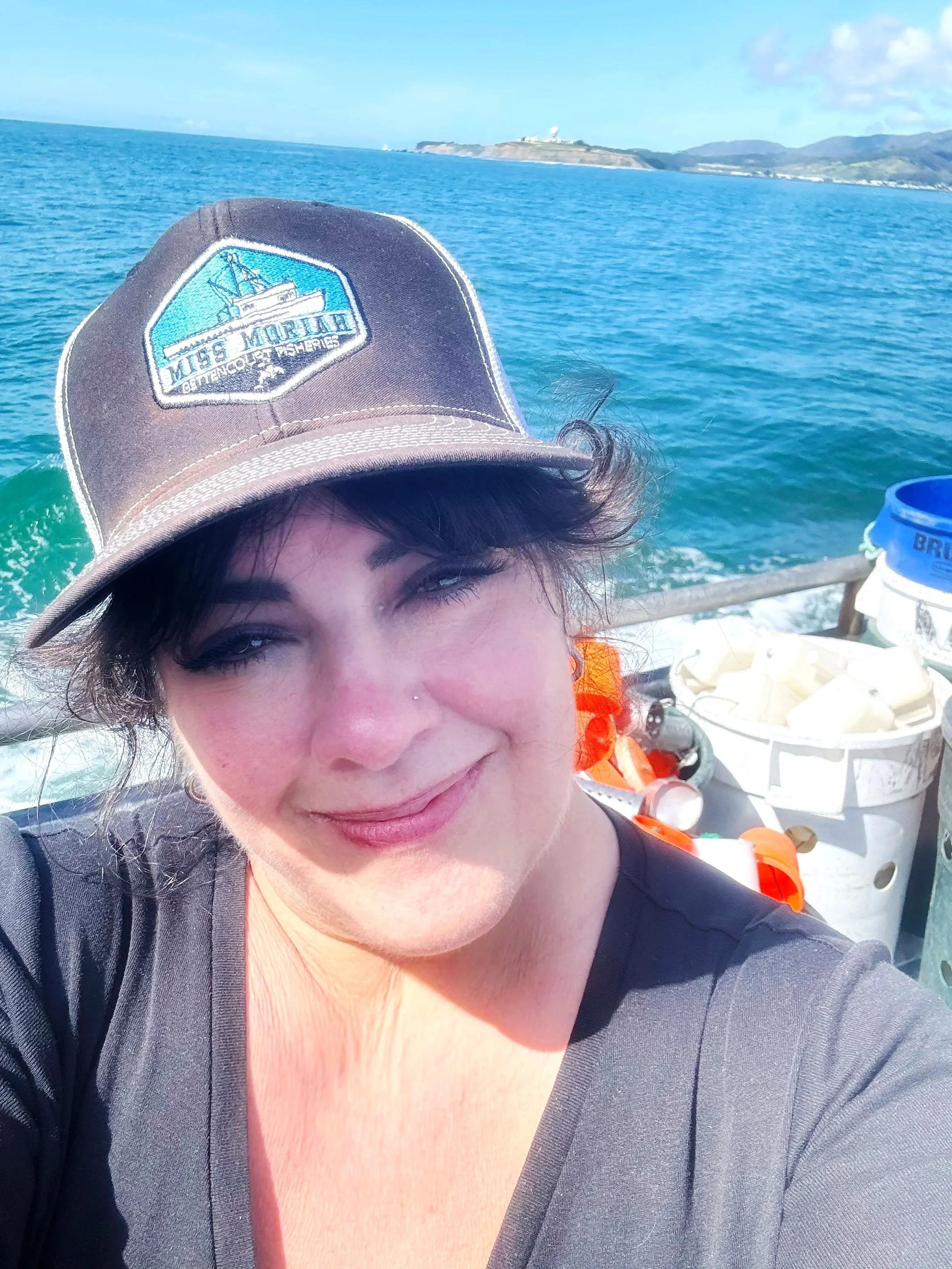 Lisa Damrosch on a boat, smiling, wearing a cap with crab gear, ocean water and coastline in the background.