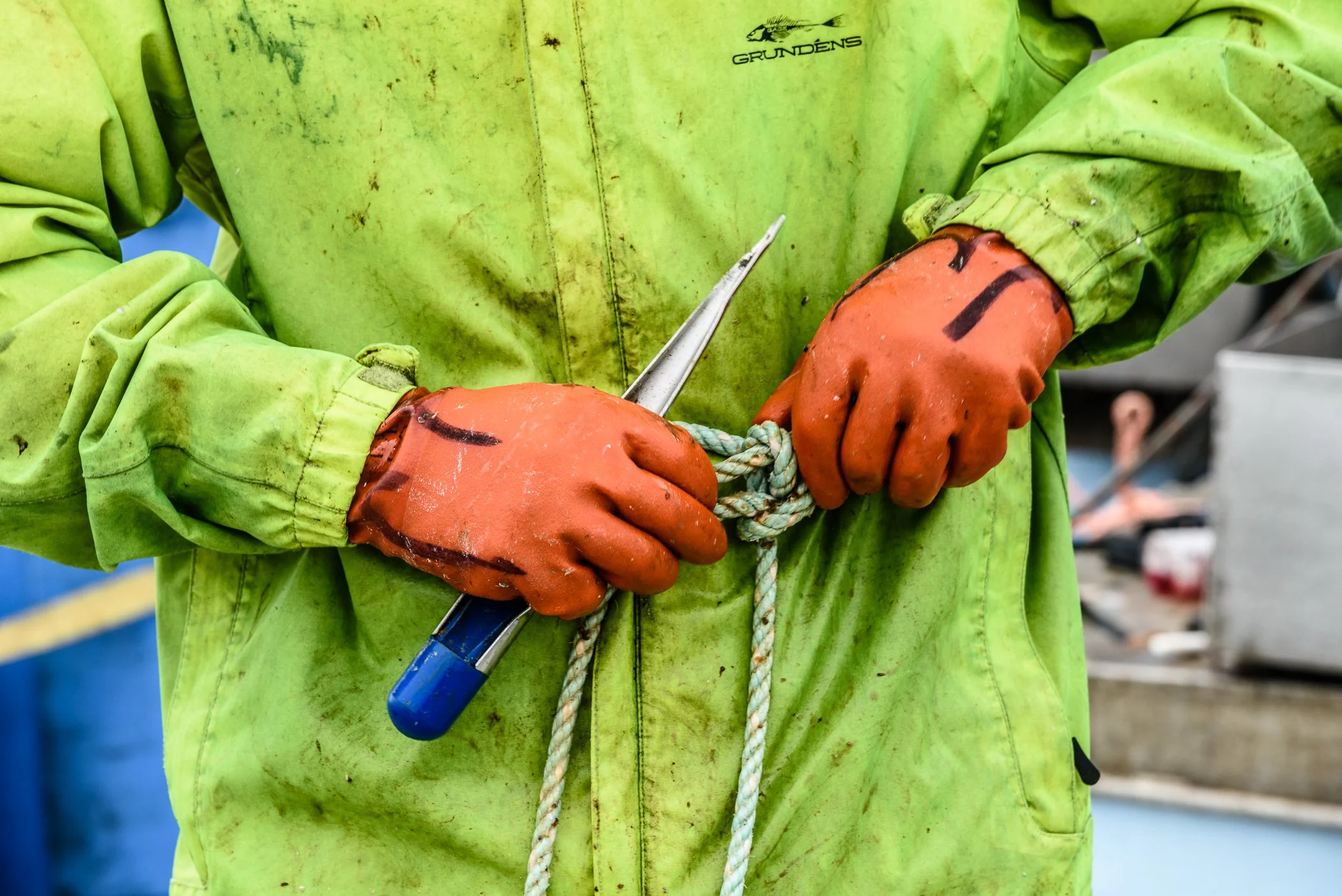 Close-up of a person wearing a green waterproof jacket and orange gloves, holding a tied rope and splicing tool k in a work environment.
