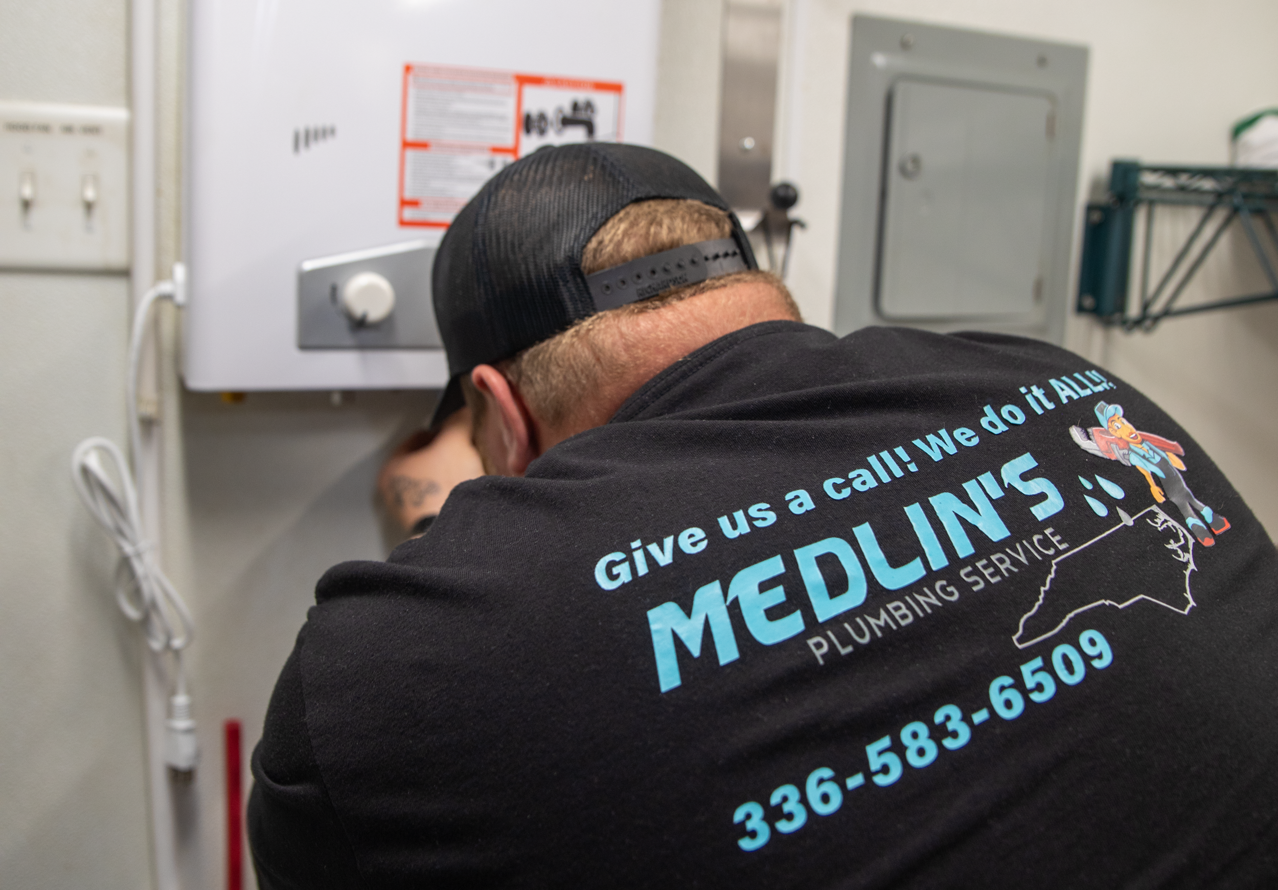 A man wearing a black baseball cap and a black T-shirt with blue and green text, bent over working on something in a utility or maintenance room. The room has electrical panels and warning notices on the wall.
