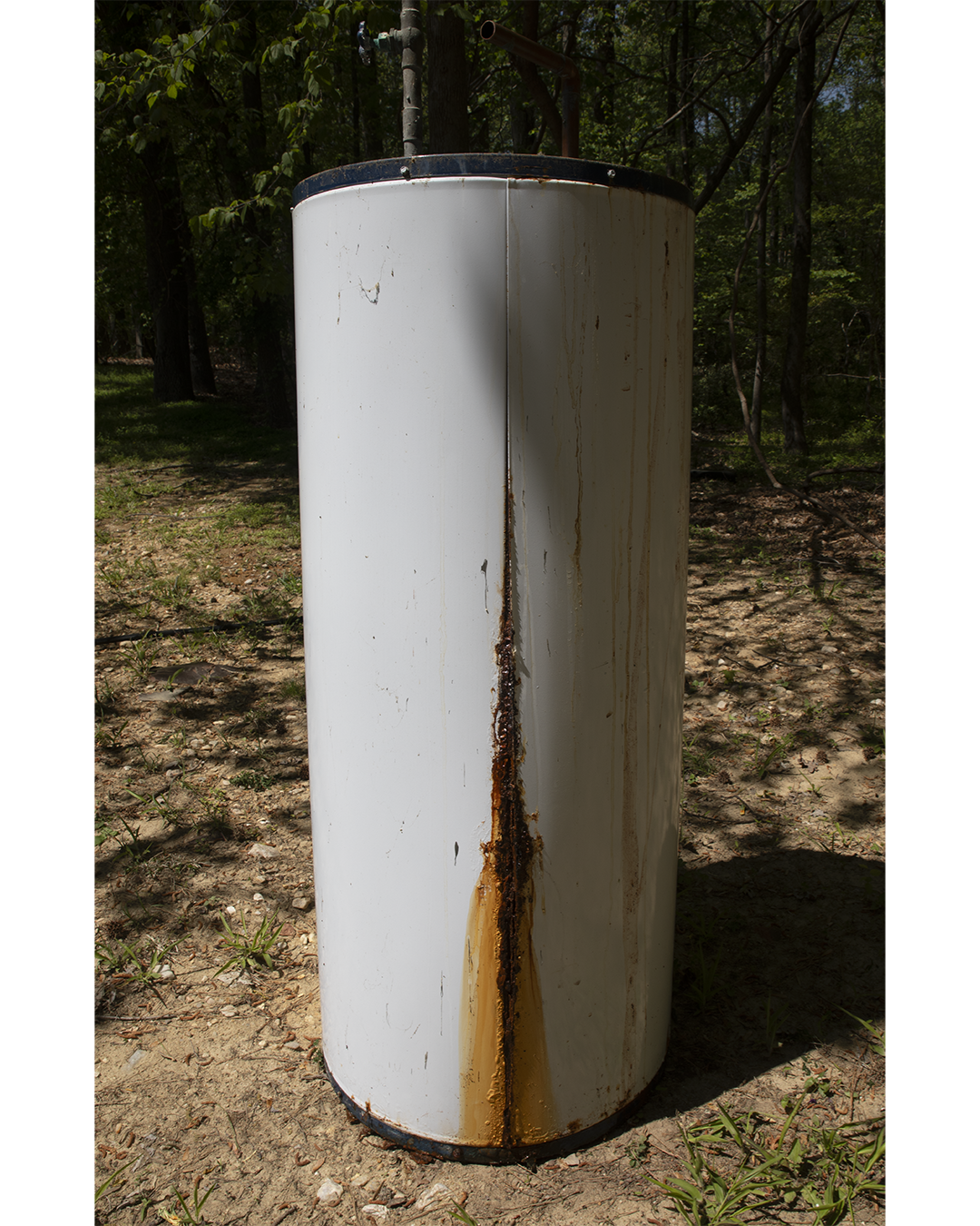 A rusty, upright, cylindrical metal tank outdoors in a wooded area. The tank has a white exterior with visible rust and corrosion, especially along a central vertical seam and near the top and bottom edges.