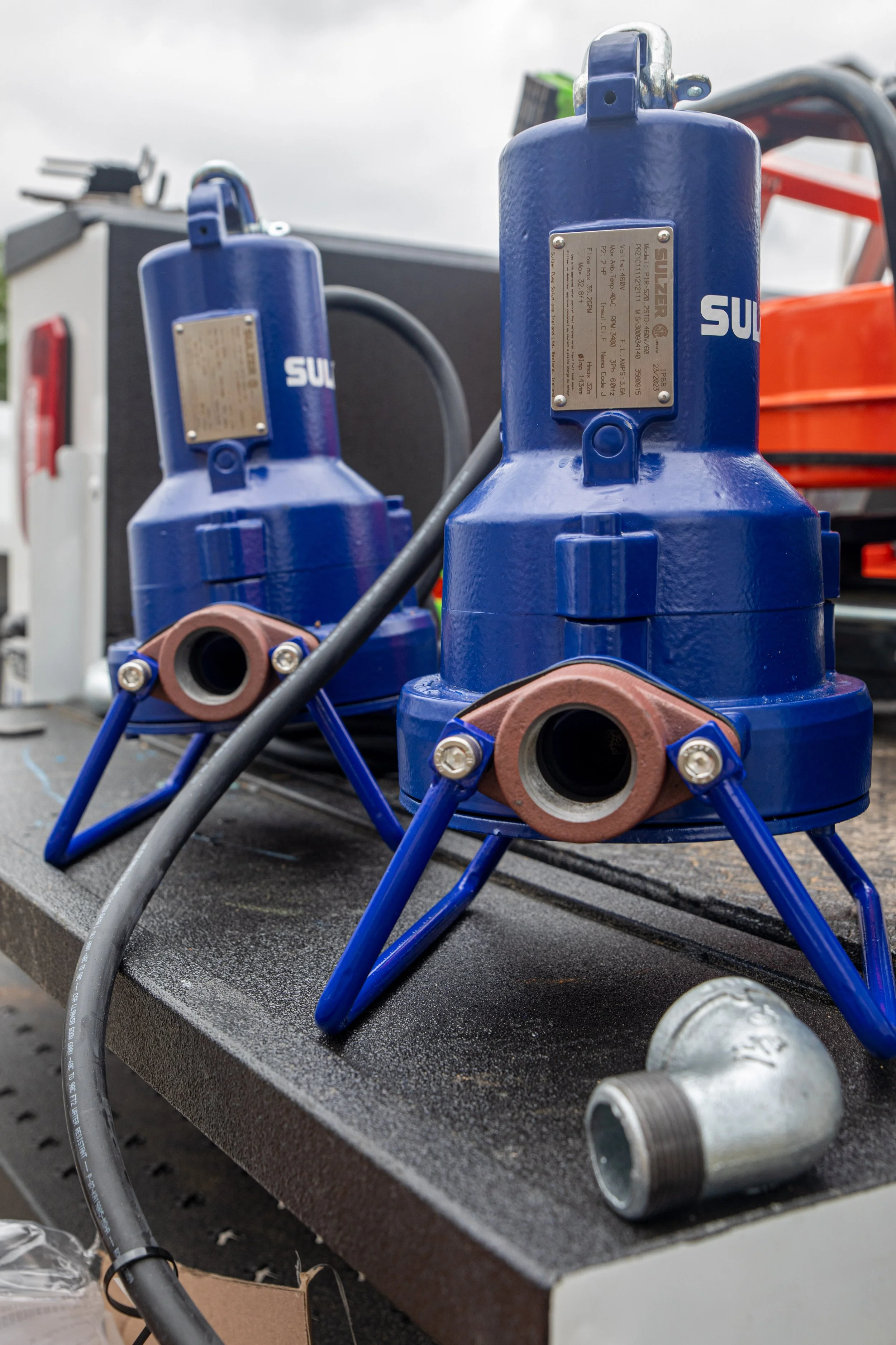 Two large blue water pumps used for car washes with hoses and labels on a workbench, with metallic fittings and a partially visible orange tool in the background.