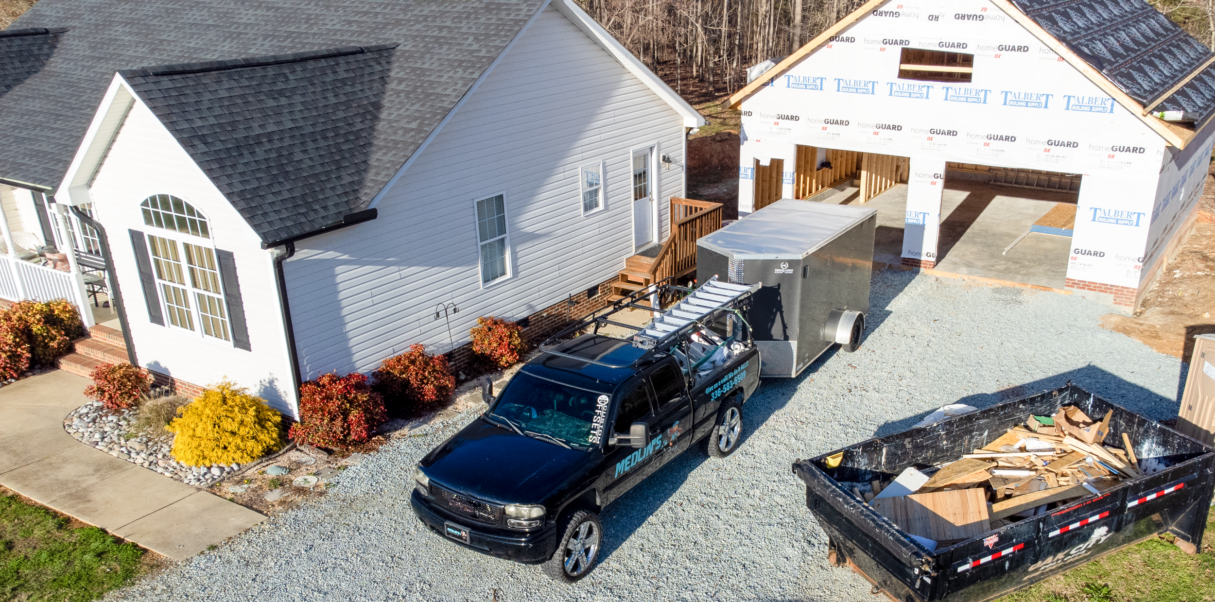 A house under construction, with one side finished and the other side with framing for a new addition, construction truck and debris in a black dumpster nearby.