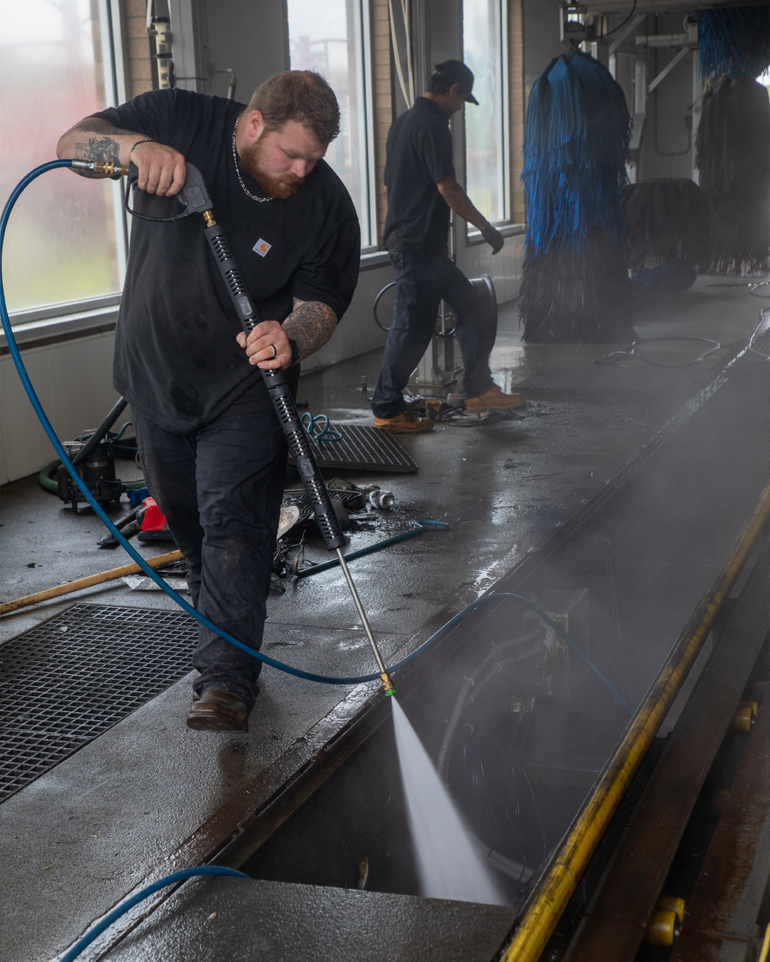 Two workers cleaning a floor drain with a power washer in an industrial or commercial building.