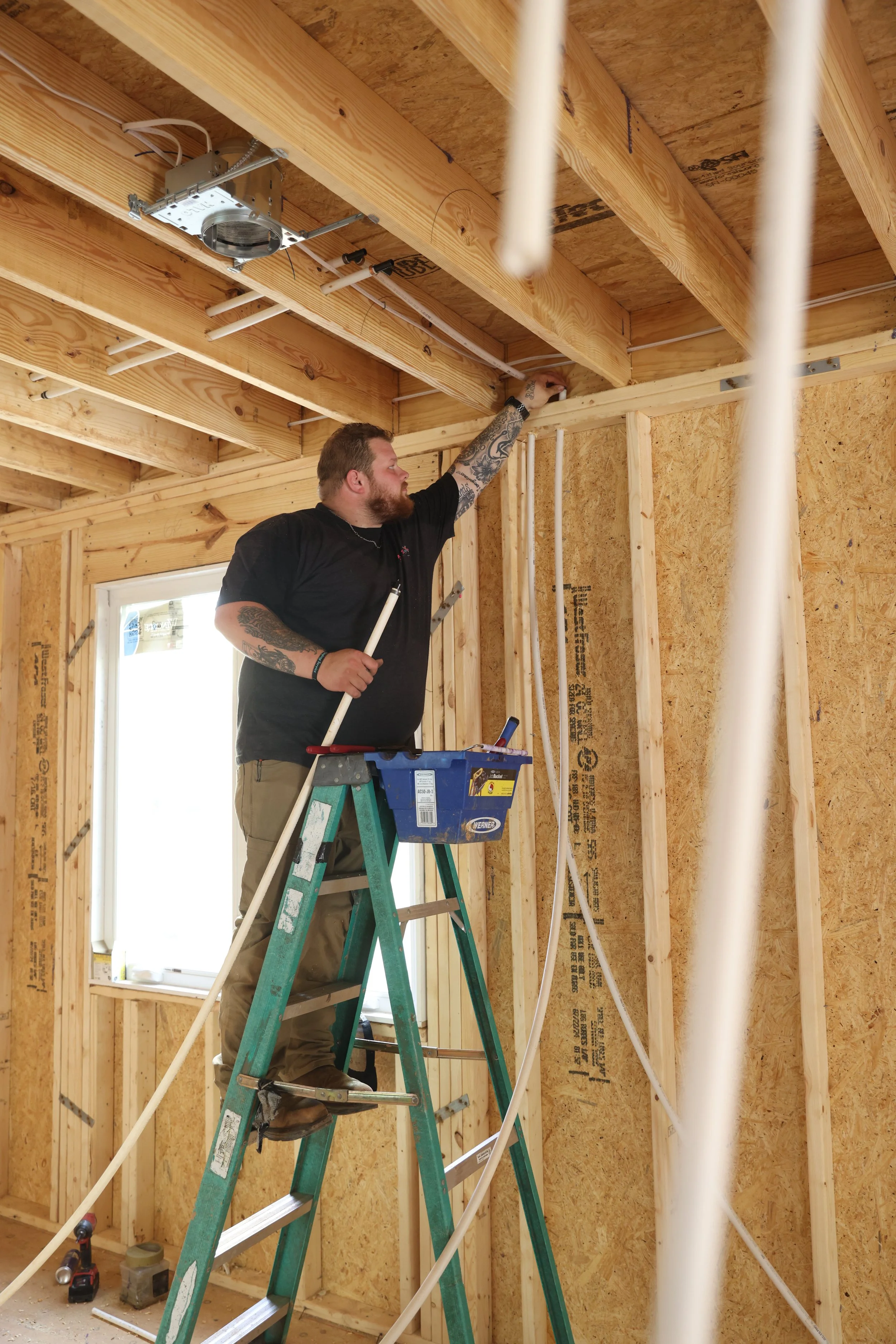 A man standing on a ladder working on the ceiling of a room under construction, surrounded by wooden framing and exposed insulation, with construction tools nearby.