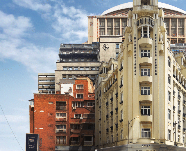A cityscape showing a mix of modern and historic buildings against a partly cloudy sky, with a clock on one building and a billboard below.