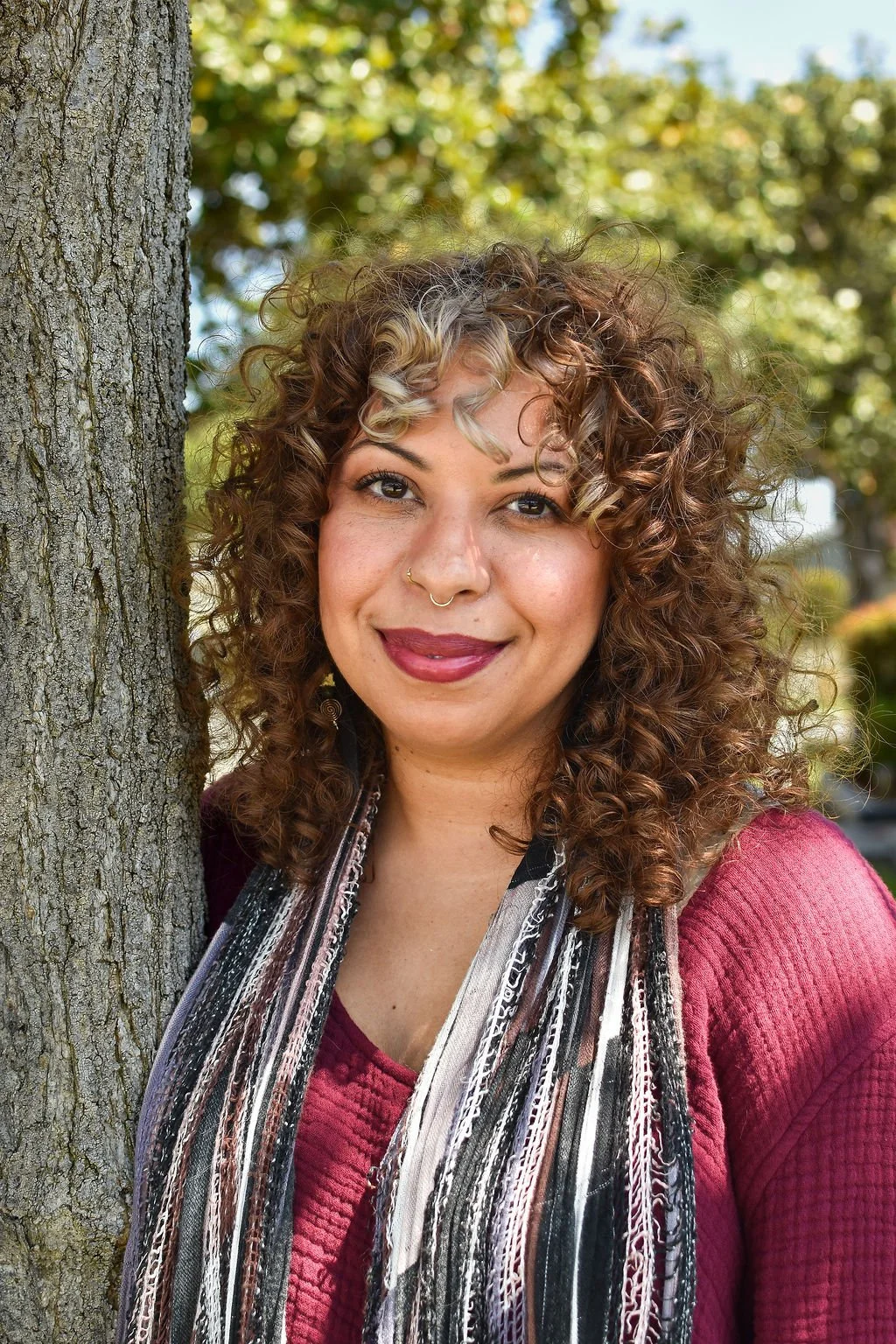 A woman with curly hair standing next to a tree in a park with autumn leaves in the background.