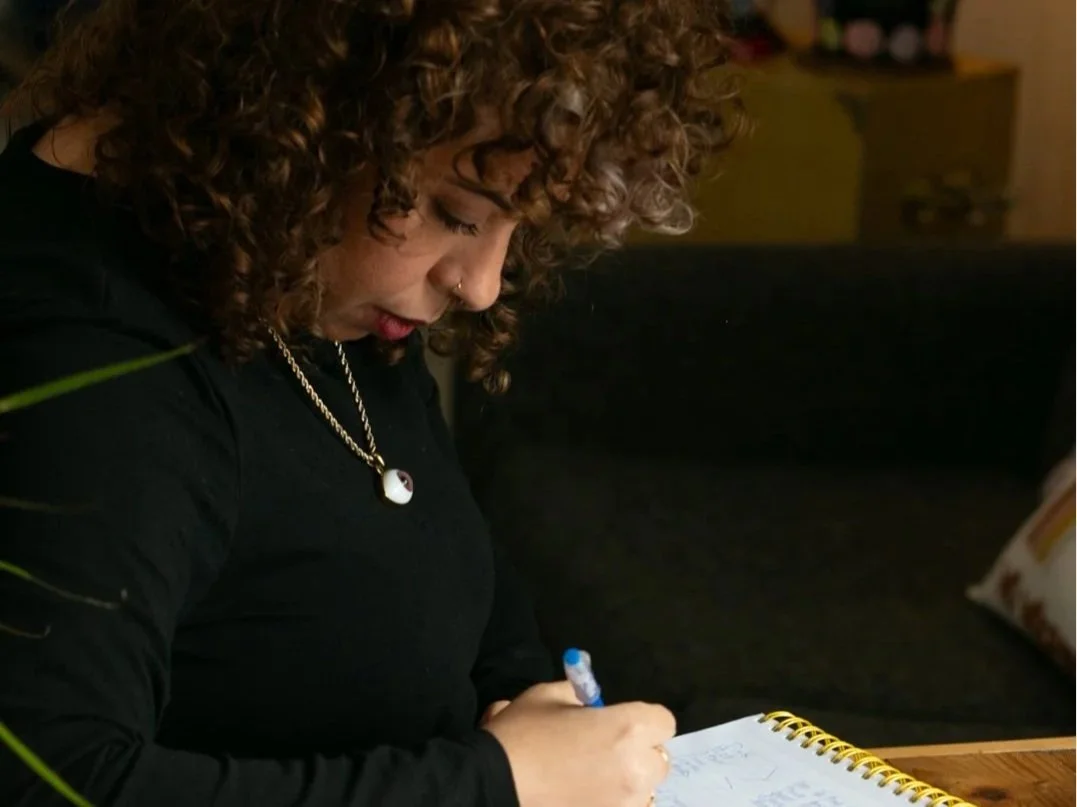 A woman with curly hair, wearing a black shirt and a necklace, is writing in a notebook with a blue pen.