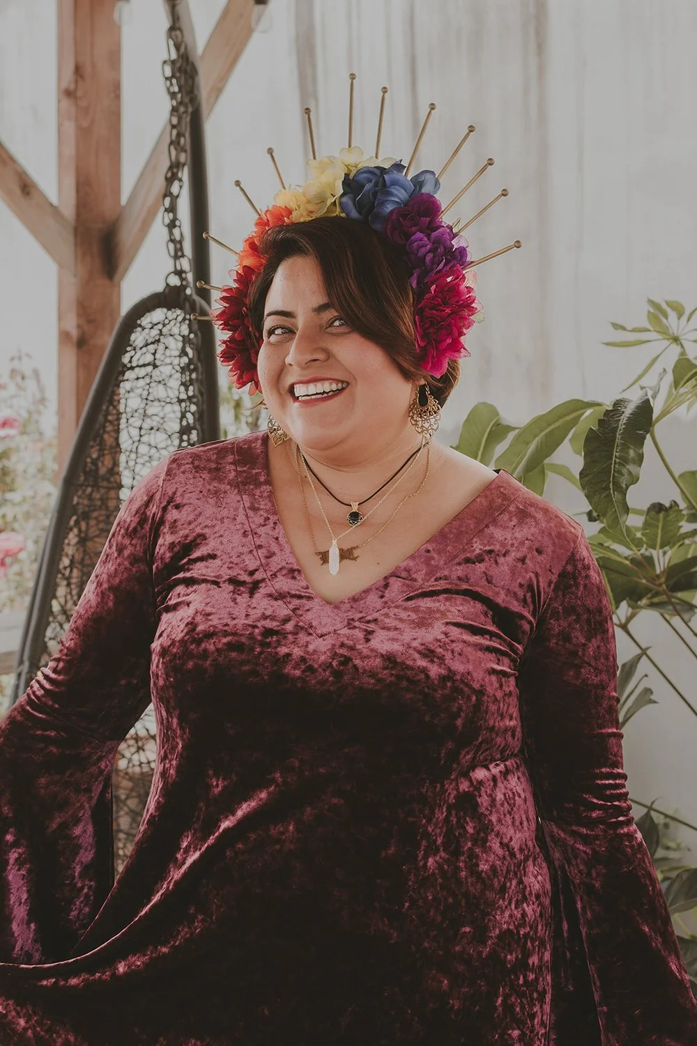 Person wearing a purple dress and colorful floral headpiece with metal spikes, smiling with plants and a wicker chair in the background.
