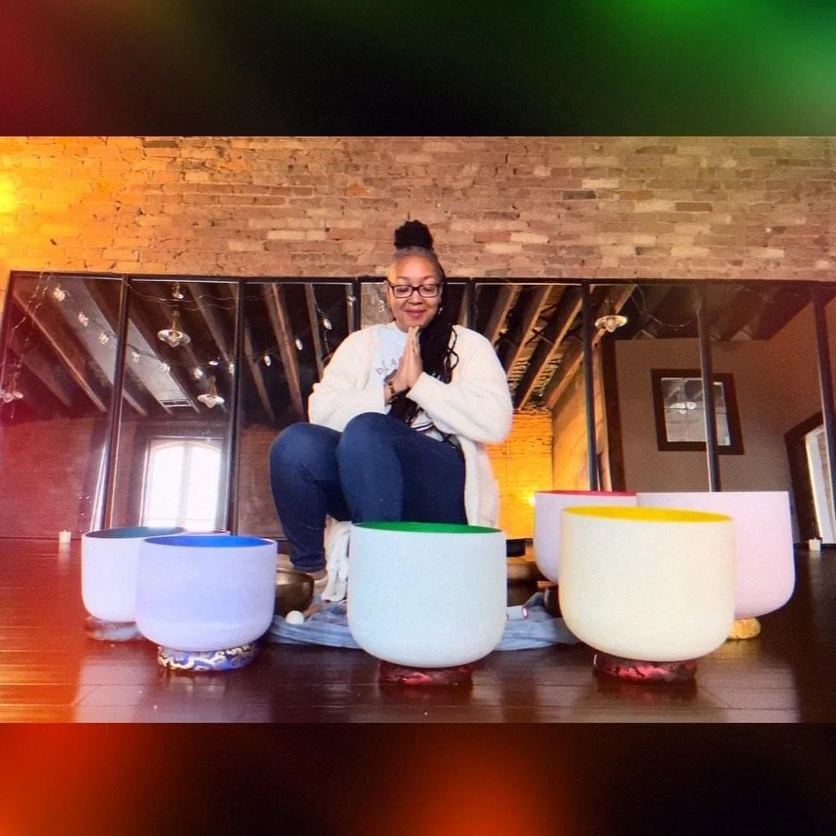 A woman sitting cross-legged with hands in prayer position, surrounded by colorful singing bowls in a room with brick walls and wooden ceiling beams.