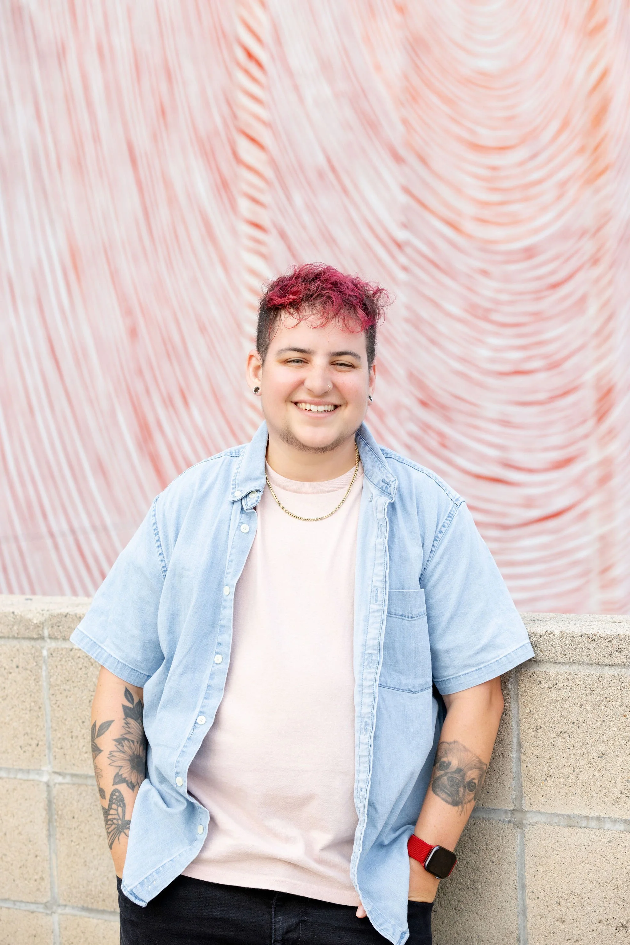 A person with short, pink hair is smiling and standing against a brick wall. They are wearing a light blue denim shirt over a white t-shirt, with tattoos on their arms and an Apple Watch with a red strap. The background features a textured, wavy red and white pattern.