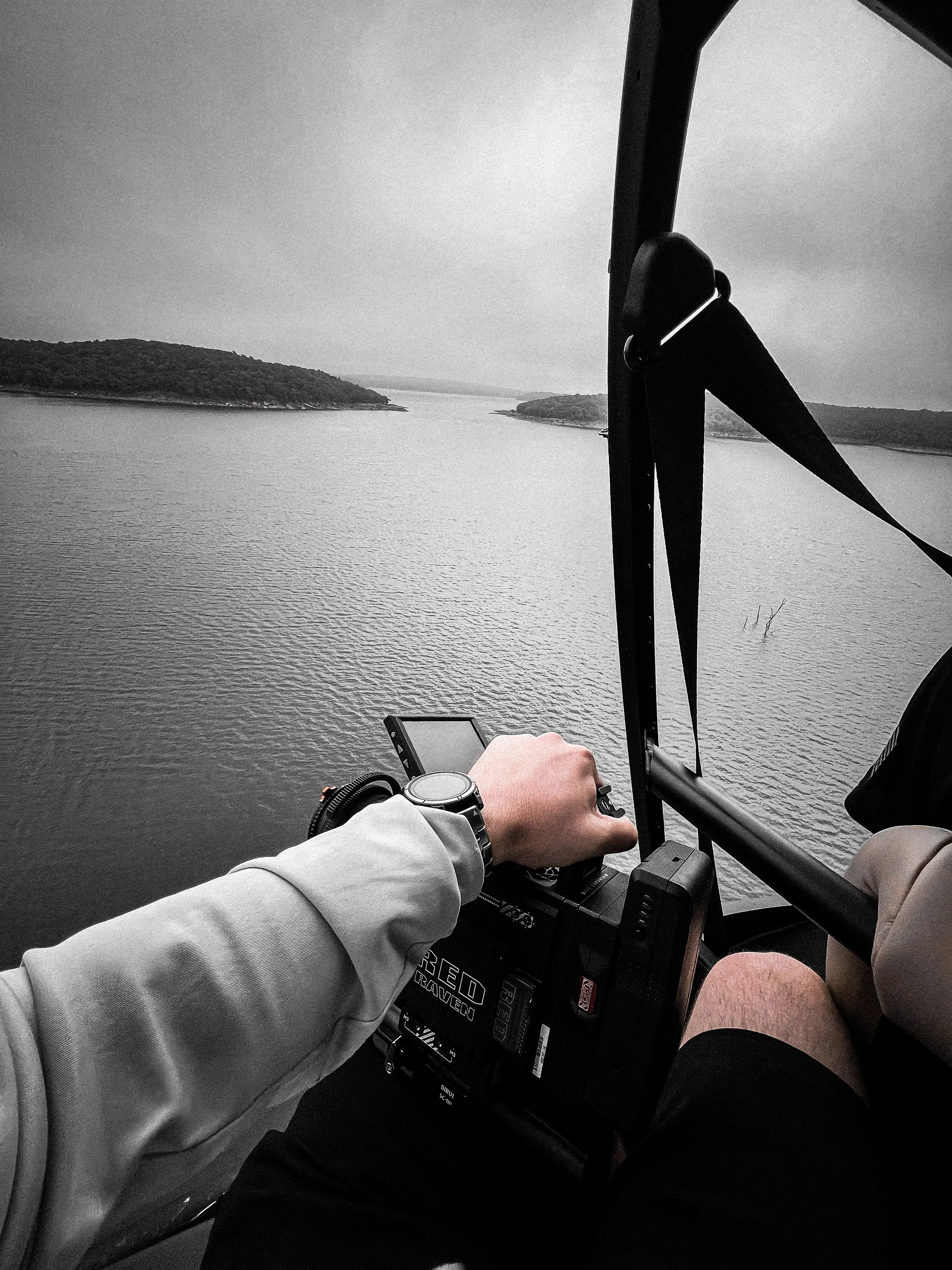 Inside a helicopter cockpit, a person wearing a watch operates the controls as they fly over a large body of water with islands in the distance, under a cloudy sky.