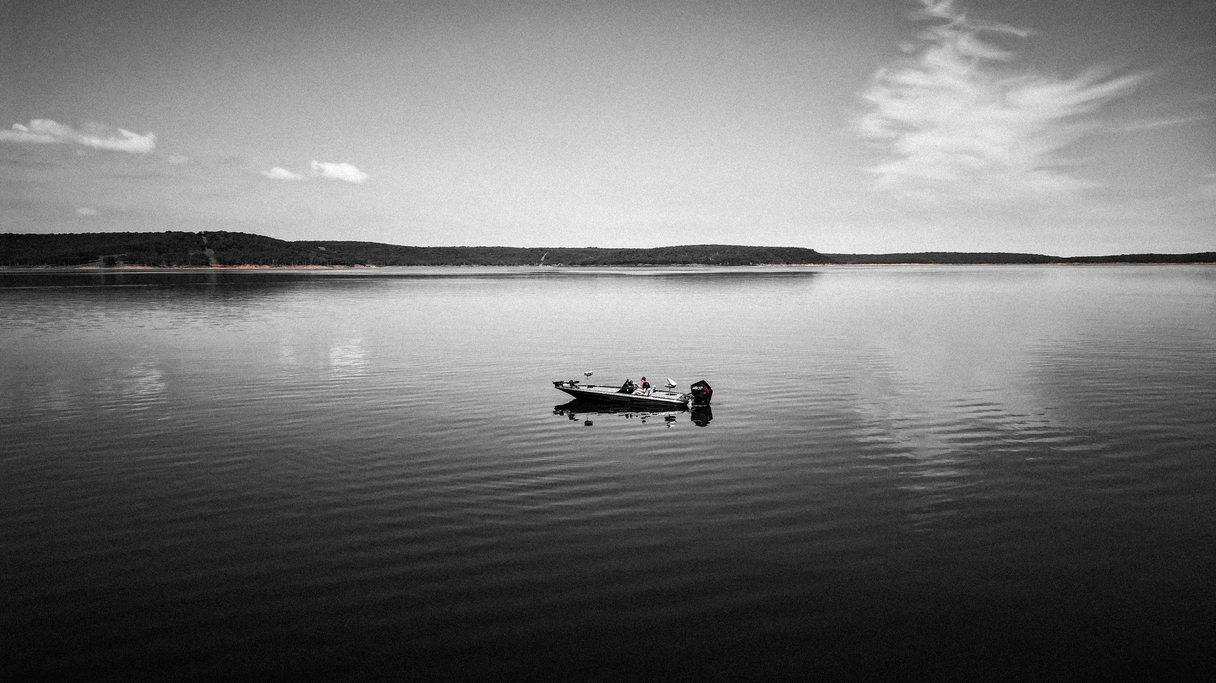 A small boat with an outboard motor floating on calm water during the day, with a distant shoreline and a partly cloudy sky in the background.