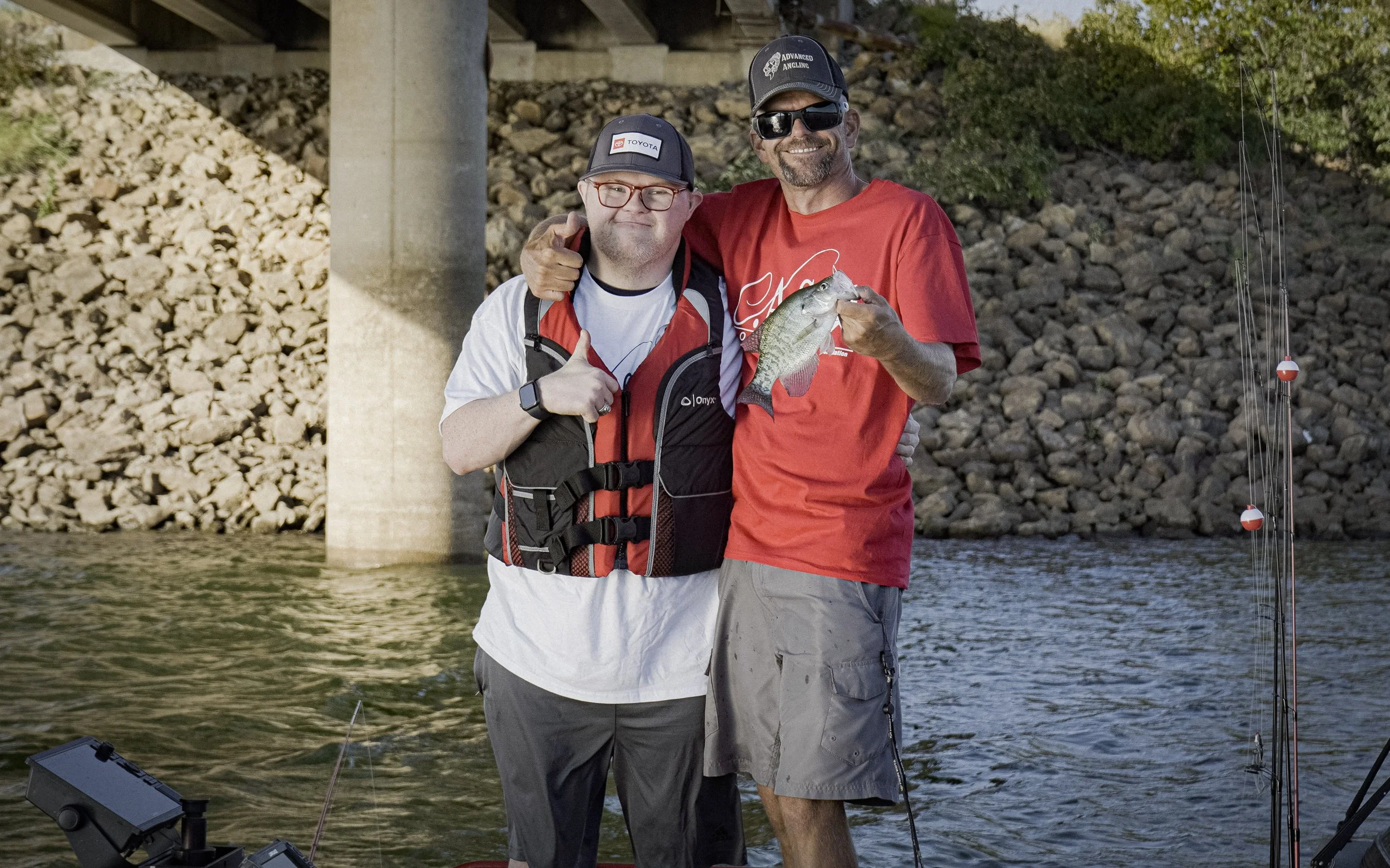 Two men standing side by side near a body of water, smiling at the camera; one holding a fish, under a bridge with rocky embankment in the background, during daytime.