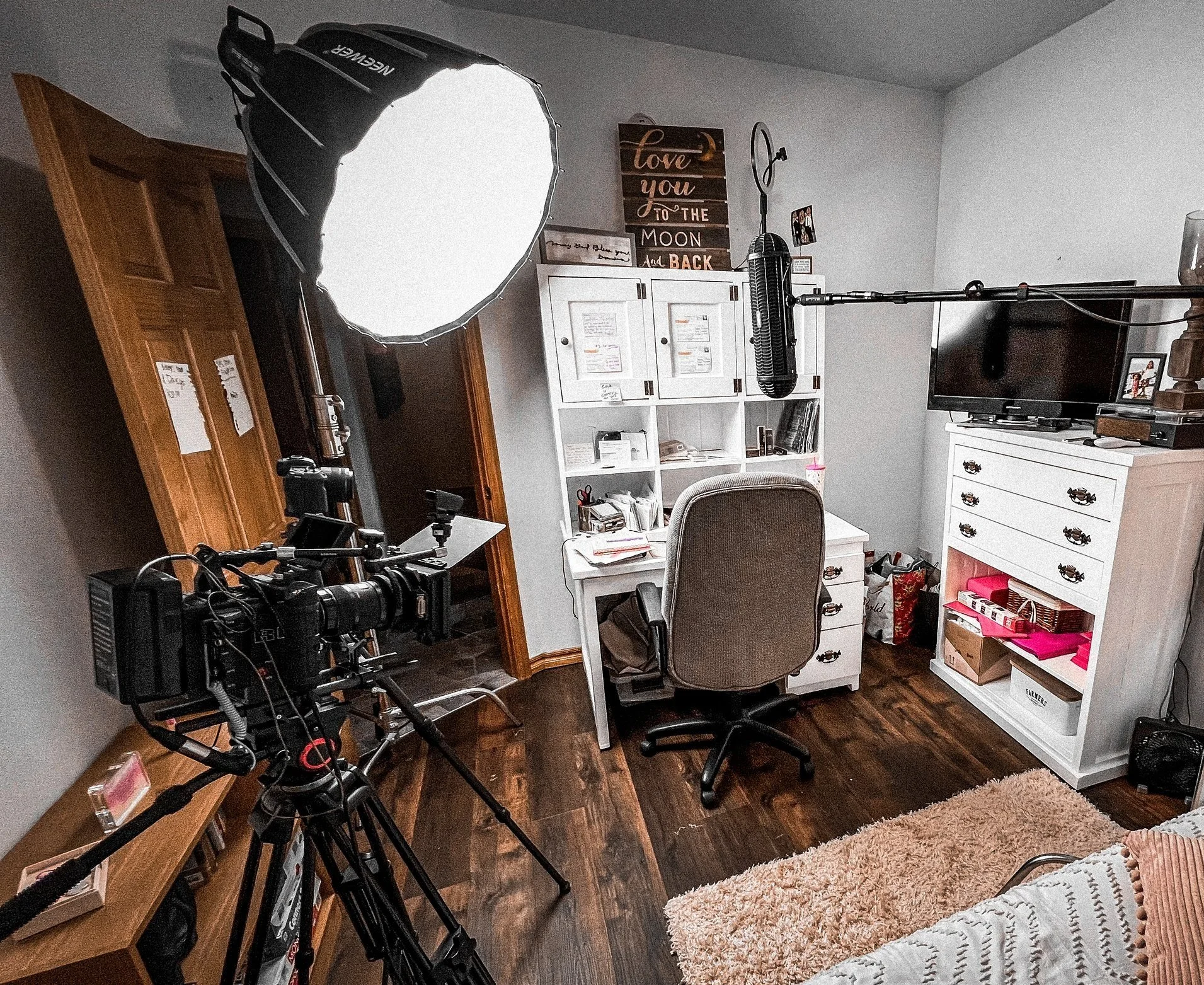 Home office setup with a white desk, gray office chair, white shelves, a flat-screen TV, and a filming camera with a large light and boom microphone in the room. There's a wooden cabinet, wall decorations, and a plush rug on the wooden floor.