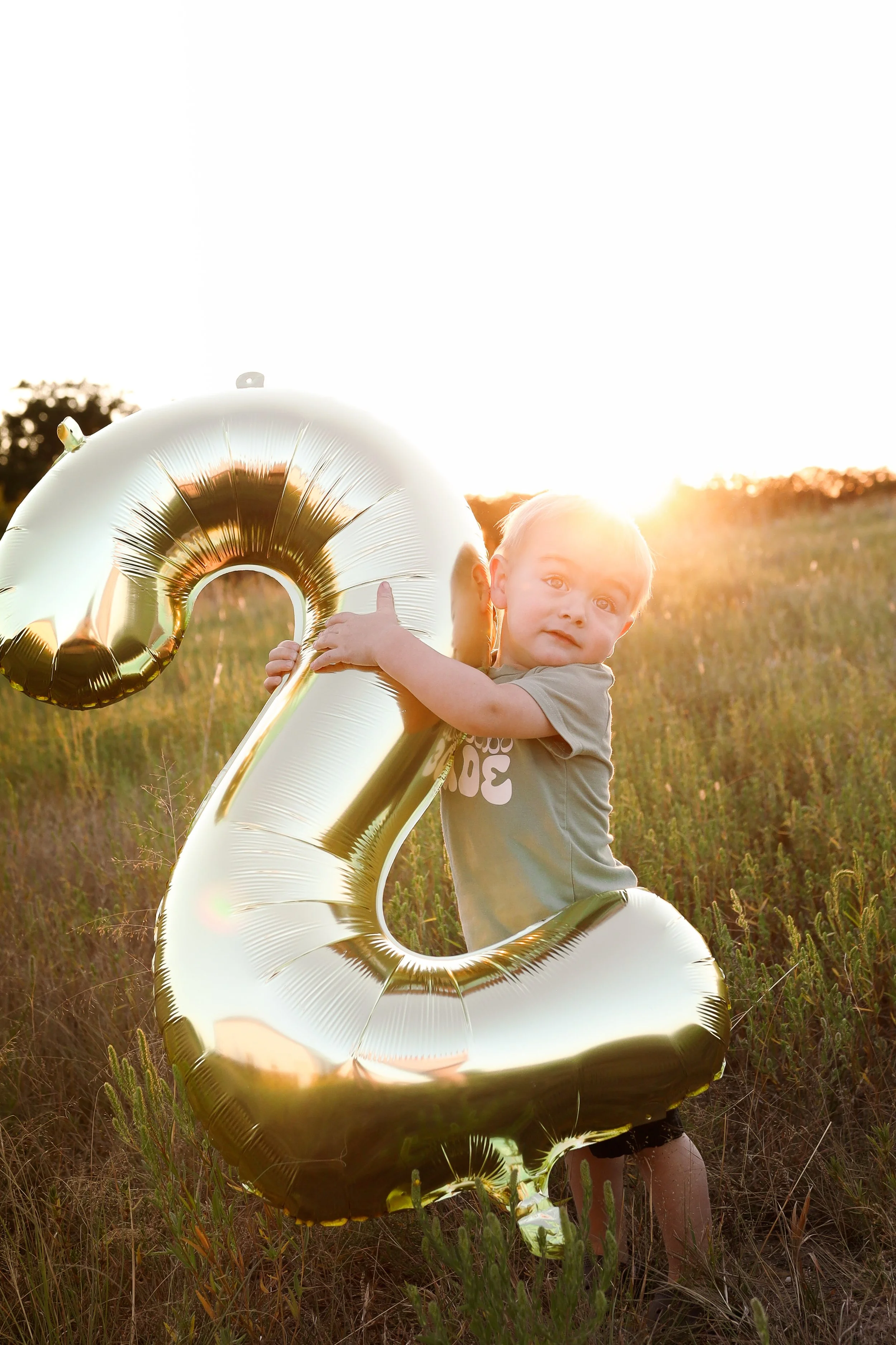 A young boy holding a large gold balloon shaped like the number 2 in a grassy field at sunset.