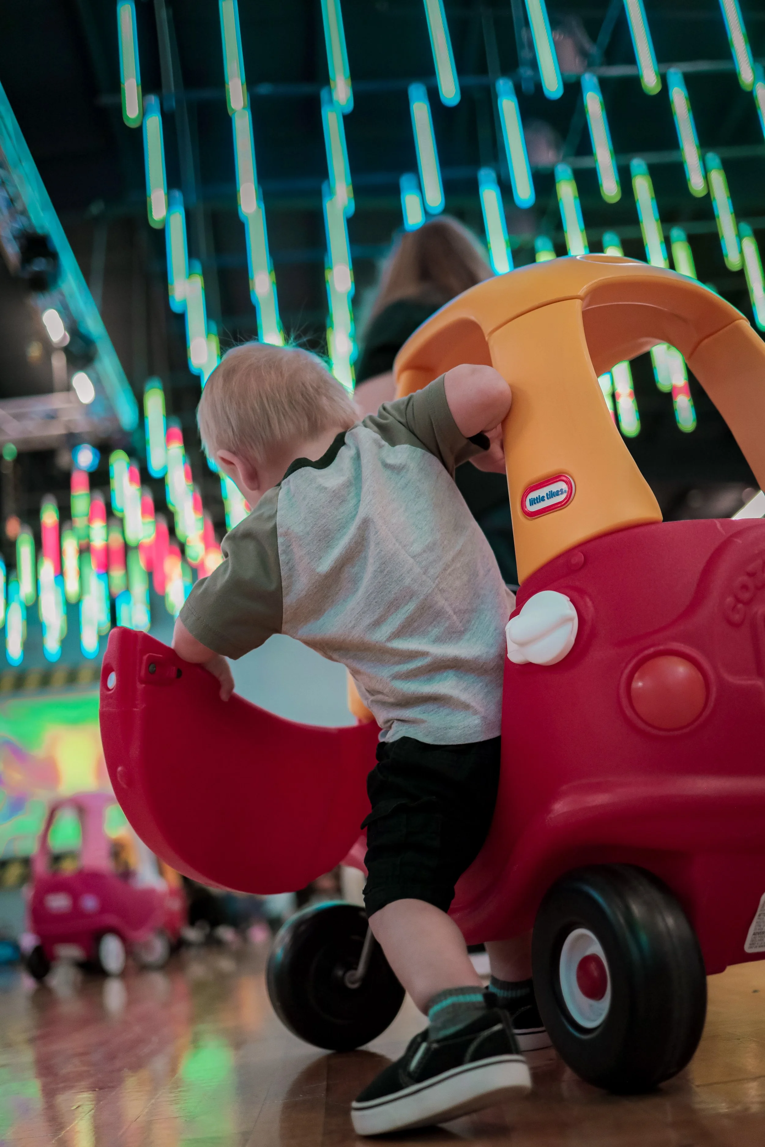 A young child climbing into a red and yellow toy vehicle at an indoor play area with colorful, illuminated decorations in the background.