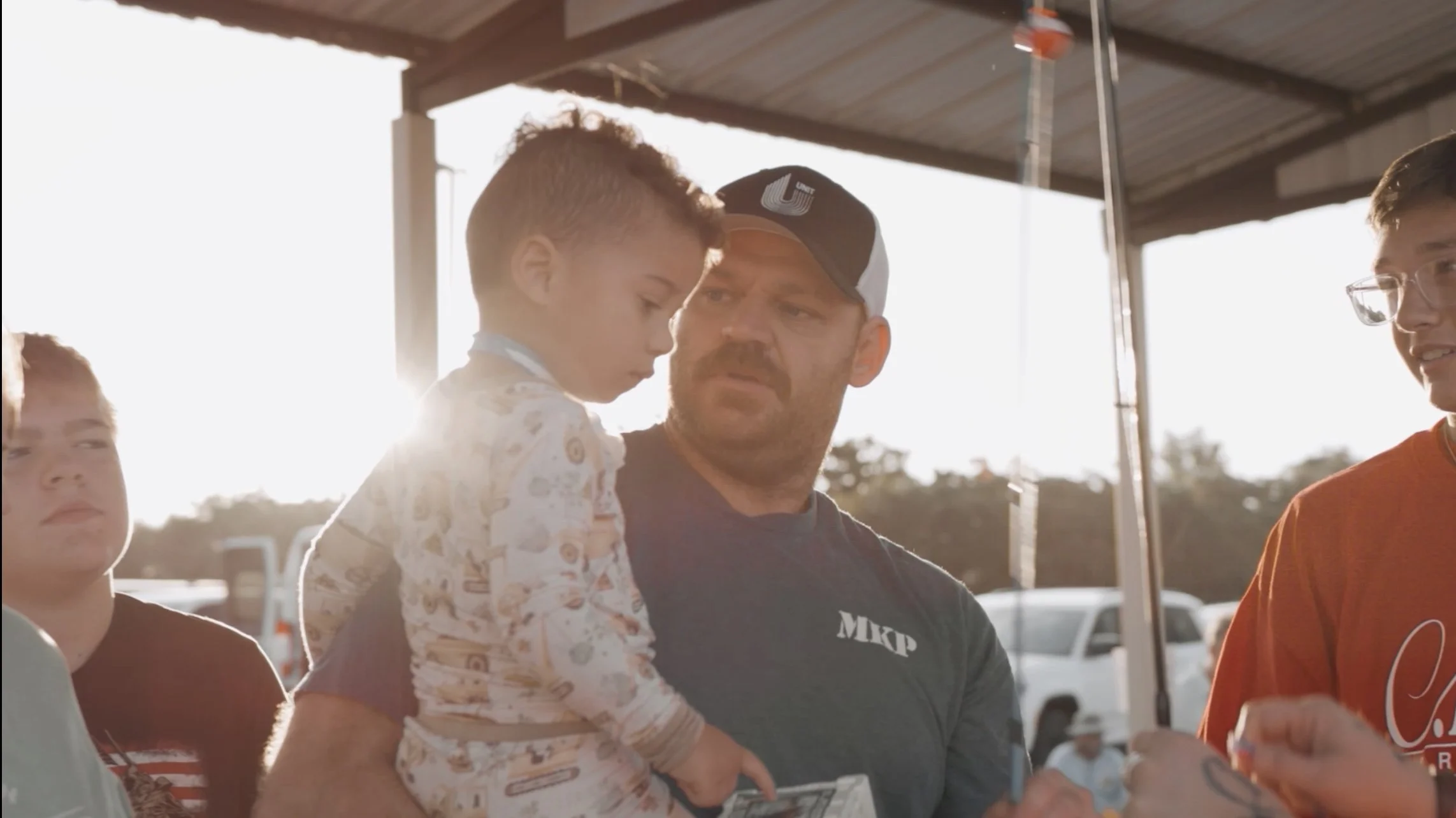 A group of men, one holding a young boy, standing under a roof with a bright background, possibly at an outdoor event or gathering.