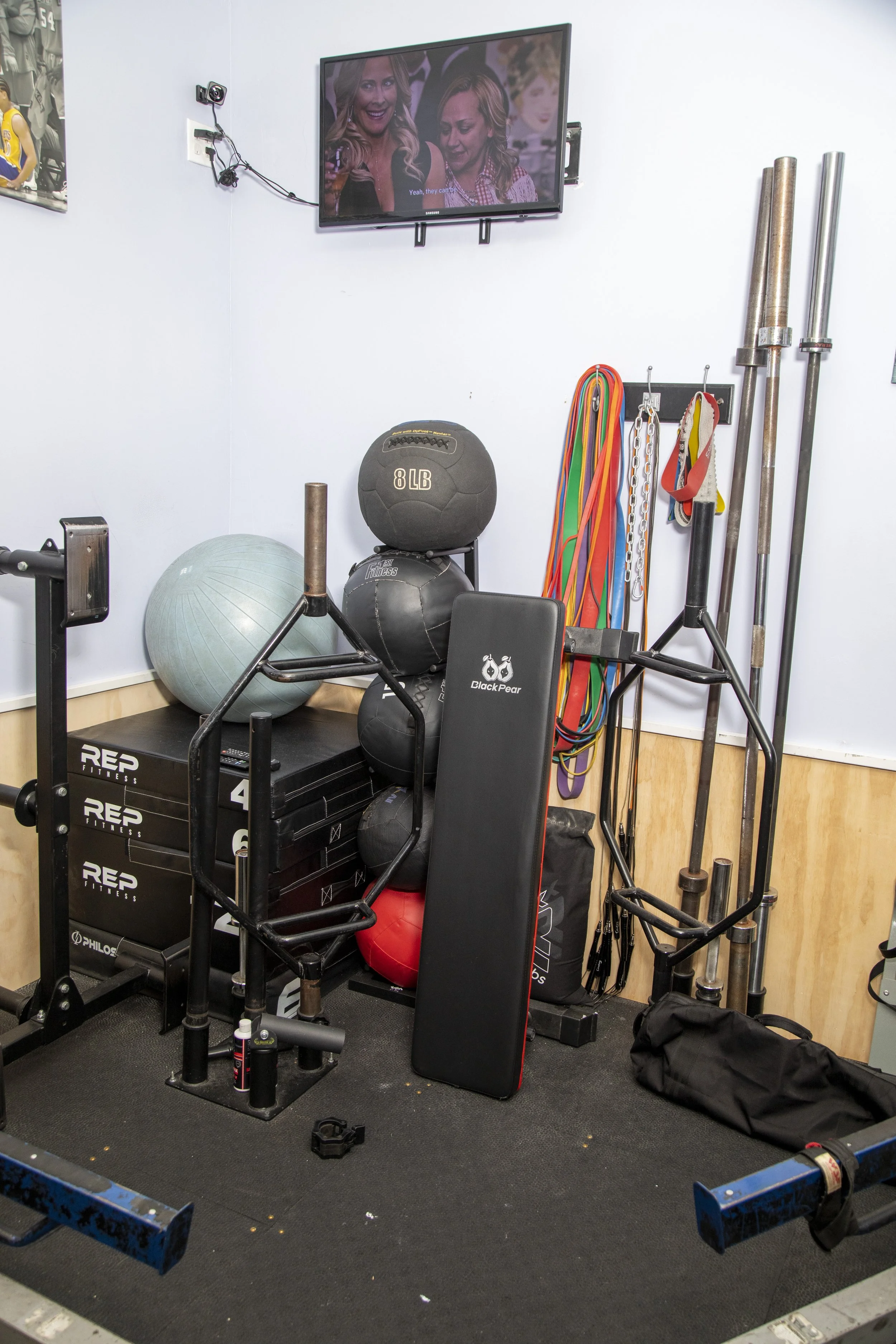 A home gym setup with various exercise equipment including a stability ball, medicine balls, resistance bands, barbells, weight plates, a punching pad, and a television mounted on the wall, displaying a scene from a show.