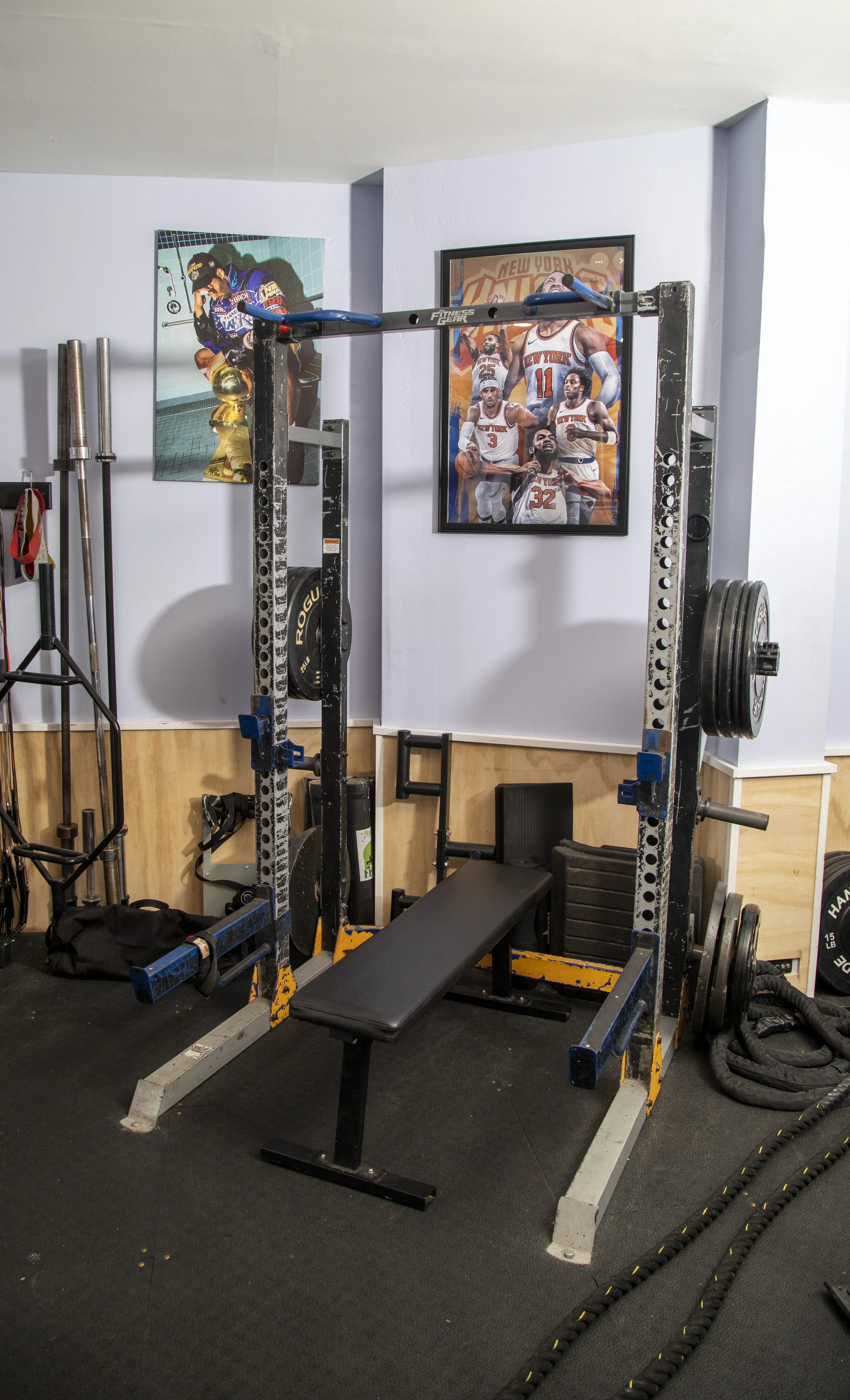 A home gym with weightlifting equipment, including a power rack with weights, a bench, and various bars. There are posters of New York Knicks basketball players on the wall.