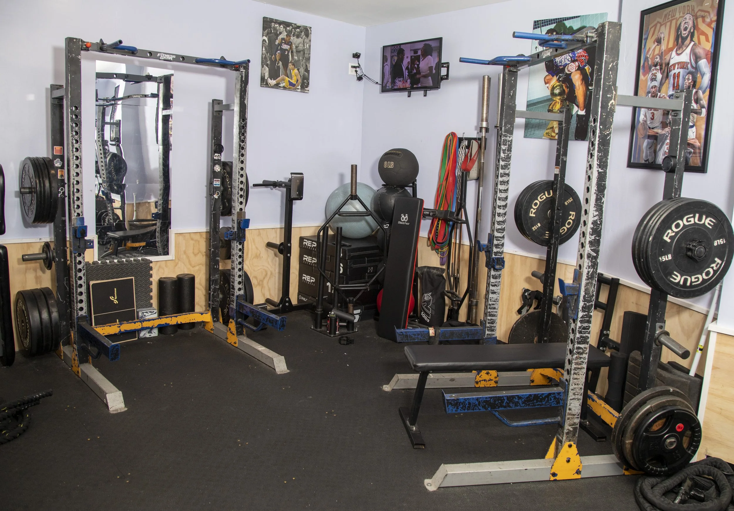 Garage gym with workout equipment including barbells, weight plates, and resistance bands. Wall decor features sports posters and a television mounted on the wall.