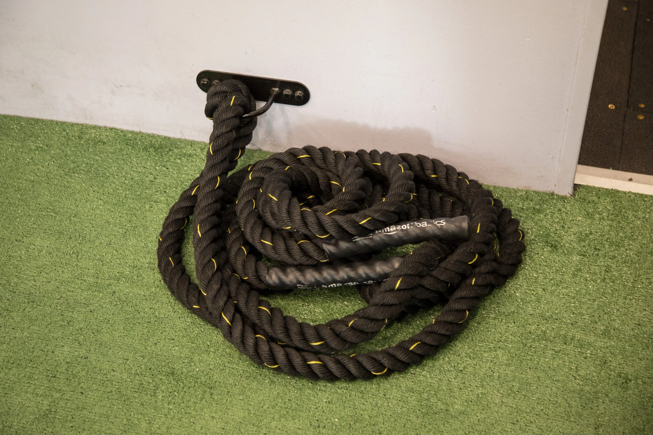 A battle rope coiled on artificial turf next to a white wall with a black attachment point.