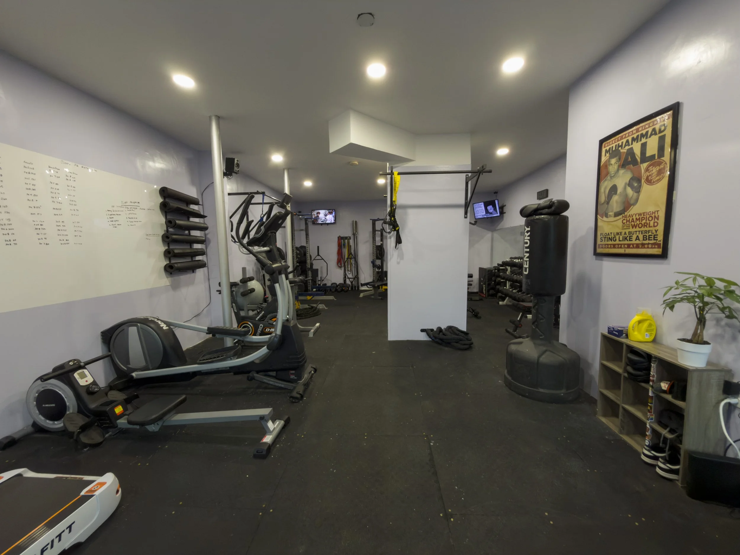 A gym with various exercise equipment, weights, and a boxing punching bag. There is a whiteboard on the left wall, small TVs mounted on the walls, and a poster of Muhammad Ali on the right wall. The floor is black with some scattered debris, and ther