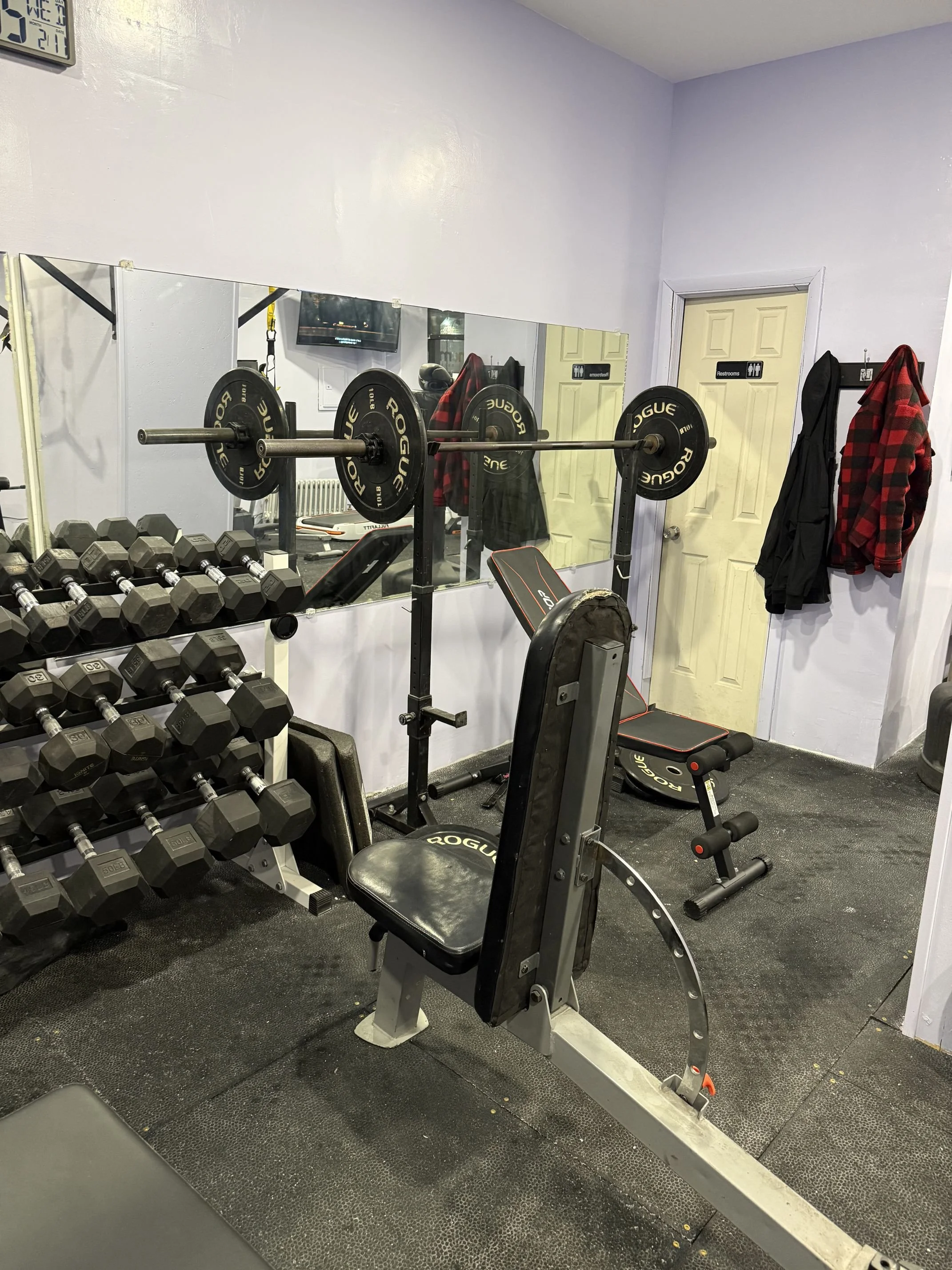 A small gym space with a weight bench, dumbbells, and barbell with weights, a mirror, coats hanging on hooks near a door, and a wall clock.
