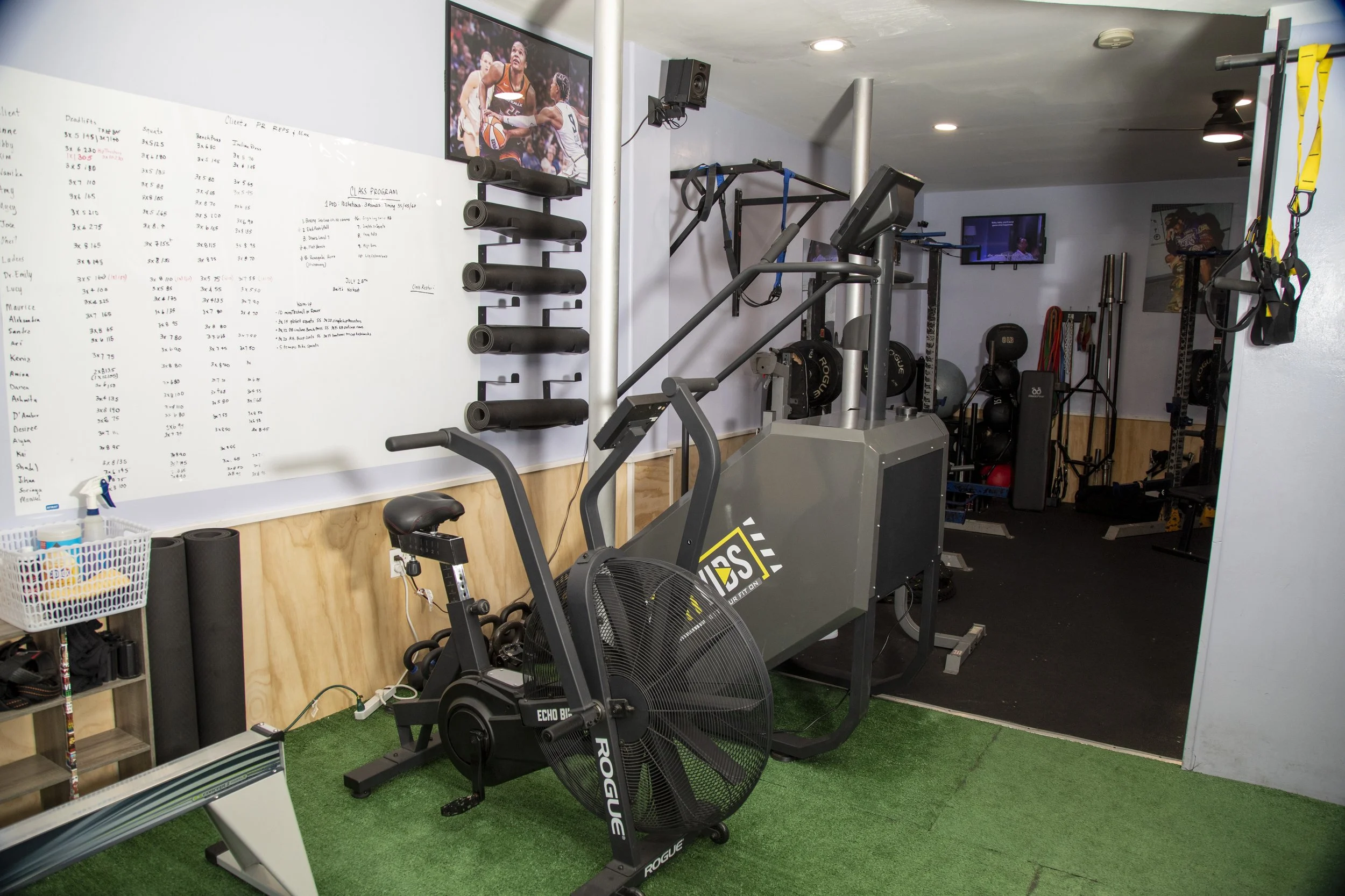 Home gym with a whiteboard, exercise equipment including a rowing machine and stationary bike, weight plates, a TV, and black and yellow suspension trainers.