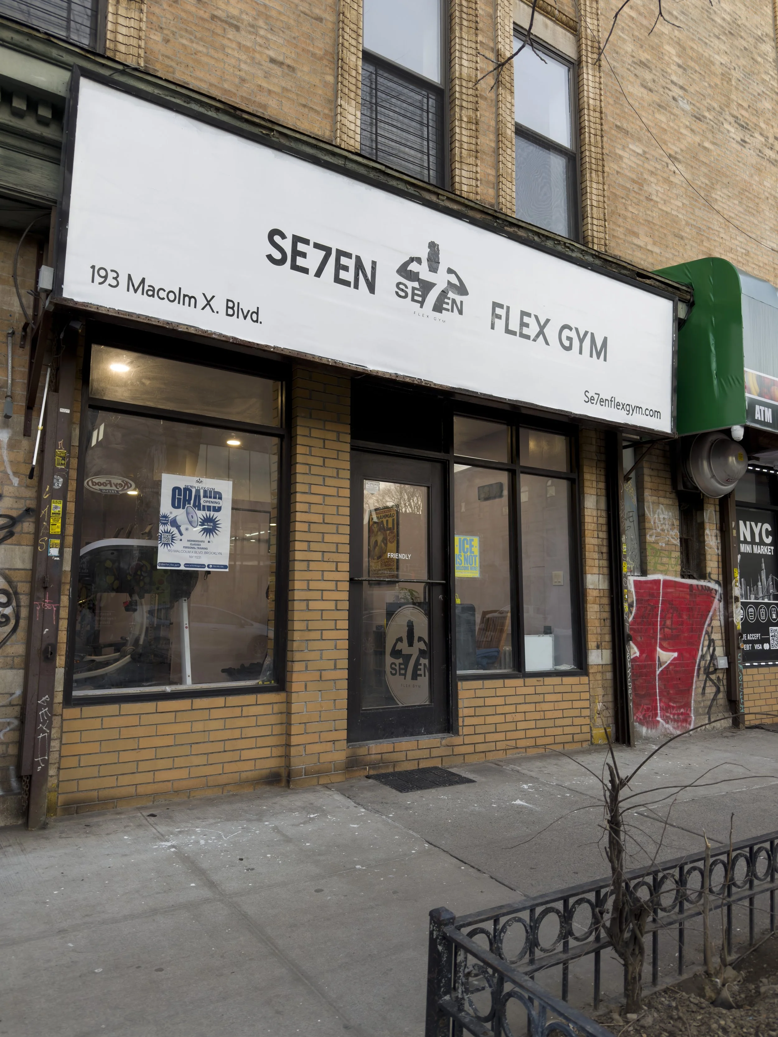 Exterior view of Se7en Flex Gym located on a city sidewalk with brick building, large window, and white sign with black text displaying the gym's name, address, and website. Inside the gym, exercise equipment is visible through the window, and graffi