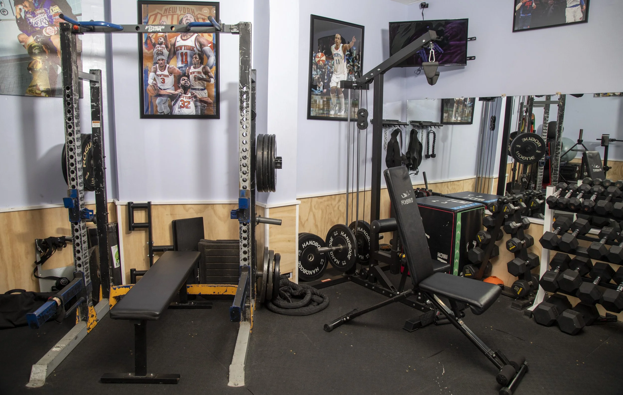 A home gym with workout equipment including a squat rack with weight plates, dumbbells, a bench, and a rowing machine. The walls feature posters of basketball players and a mirror, with a TV mounted on the wall.