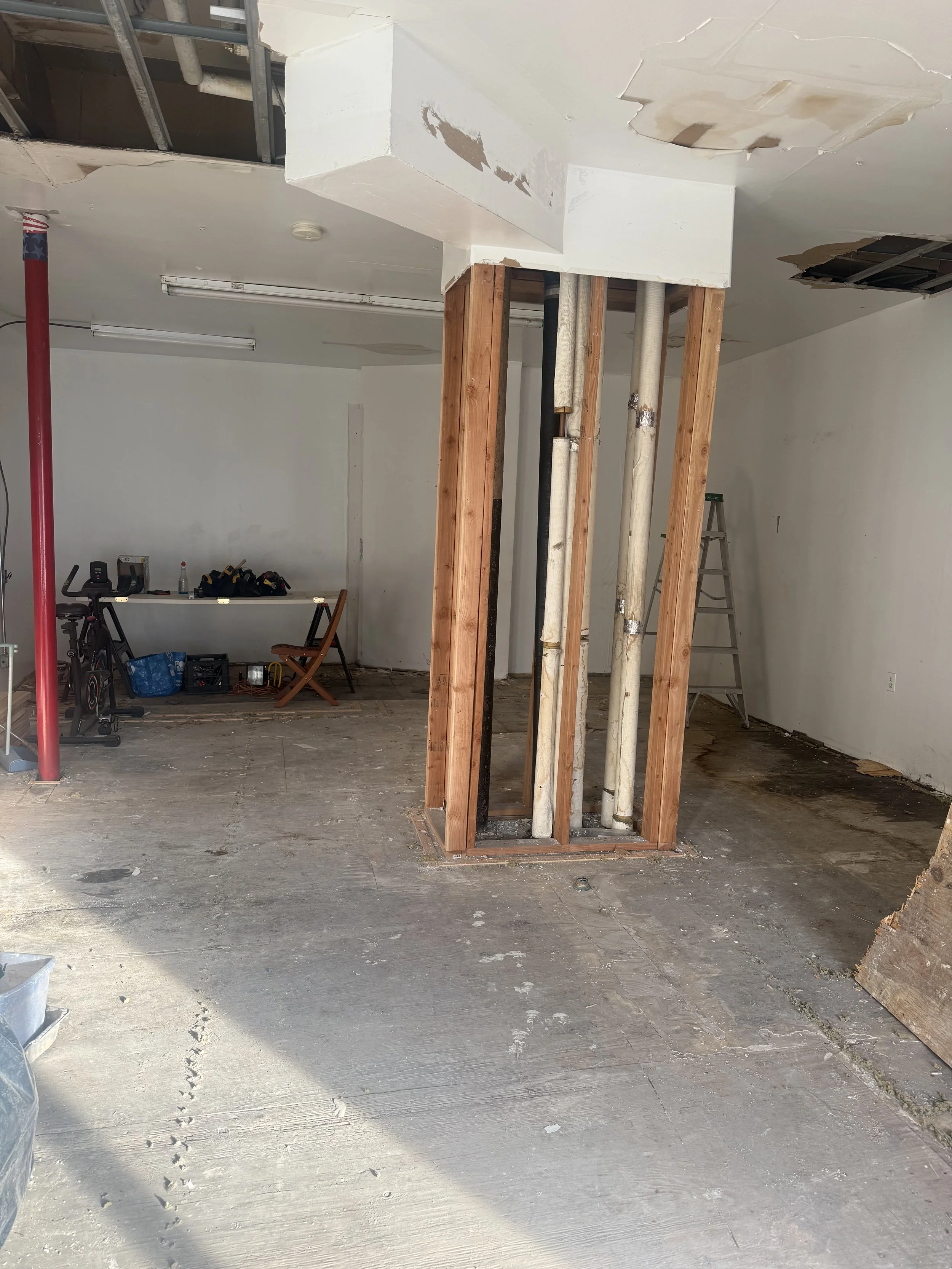 Interior view of a building under renovation with exposed pipes and wooden framing, unfinished ceiling, and construction tools and materials scattered around.