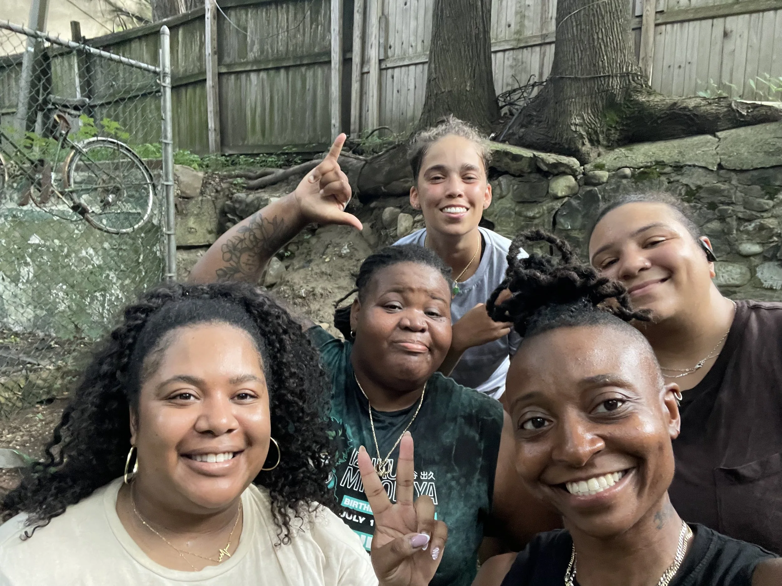 Six women posing outdoors for a photo, smiling, and making peace signs, with trees, a wooden fence, and a stone wall in the background.